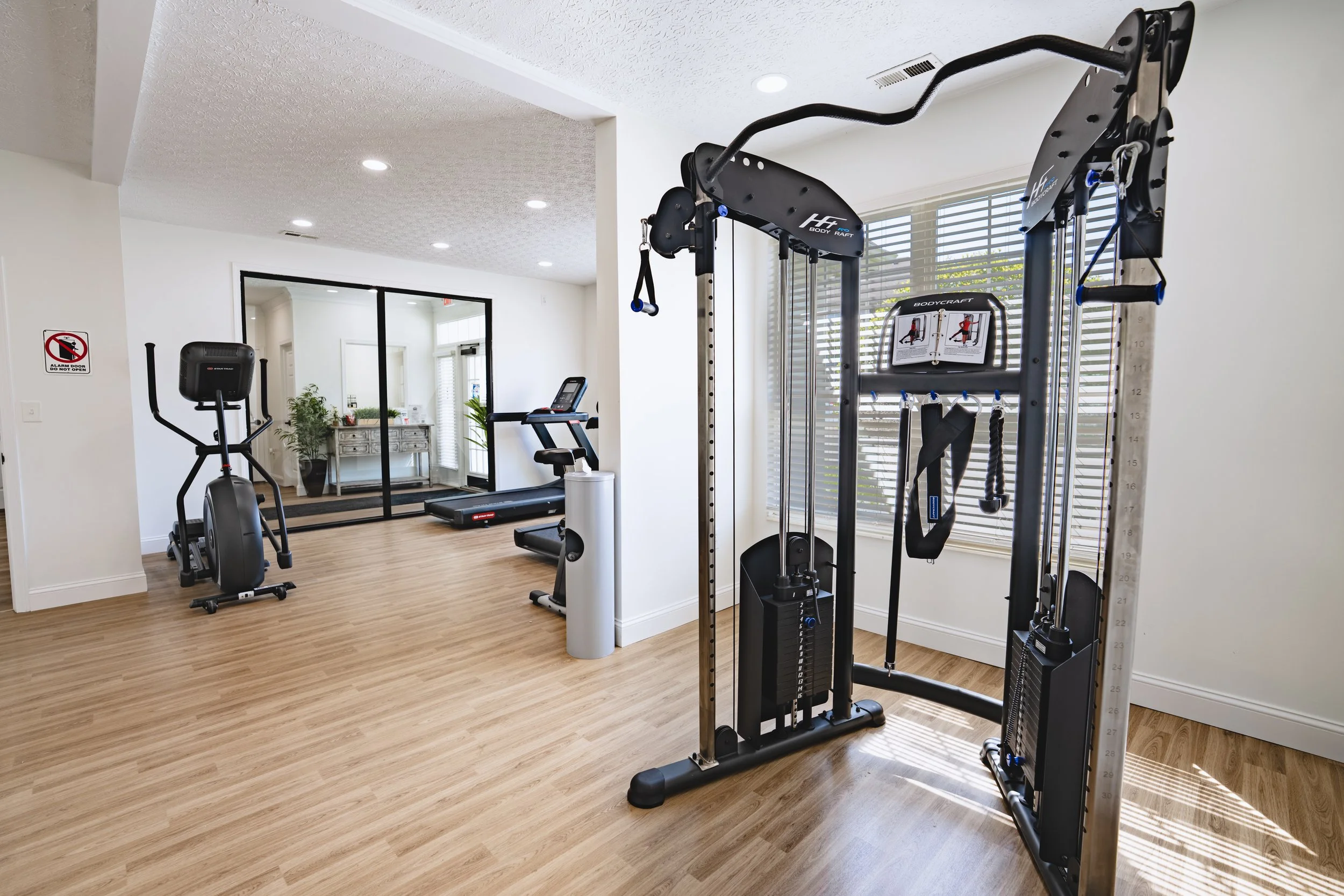 Home gym with workout equipment including a treadmill, elliptical, and a multi-function cable machine, in a bright room with hardwood floors and large window blinds.