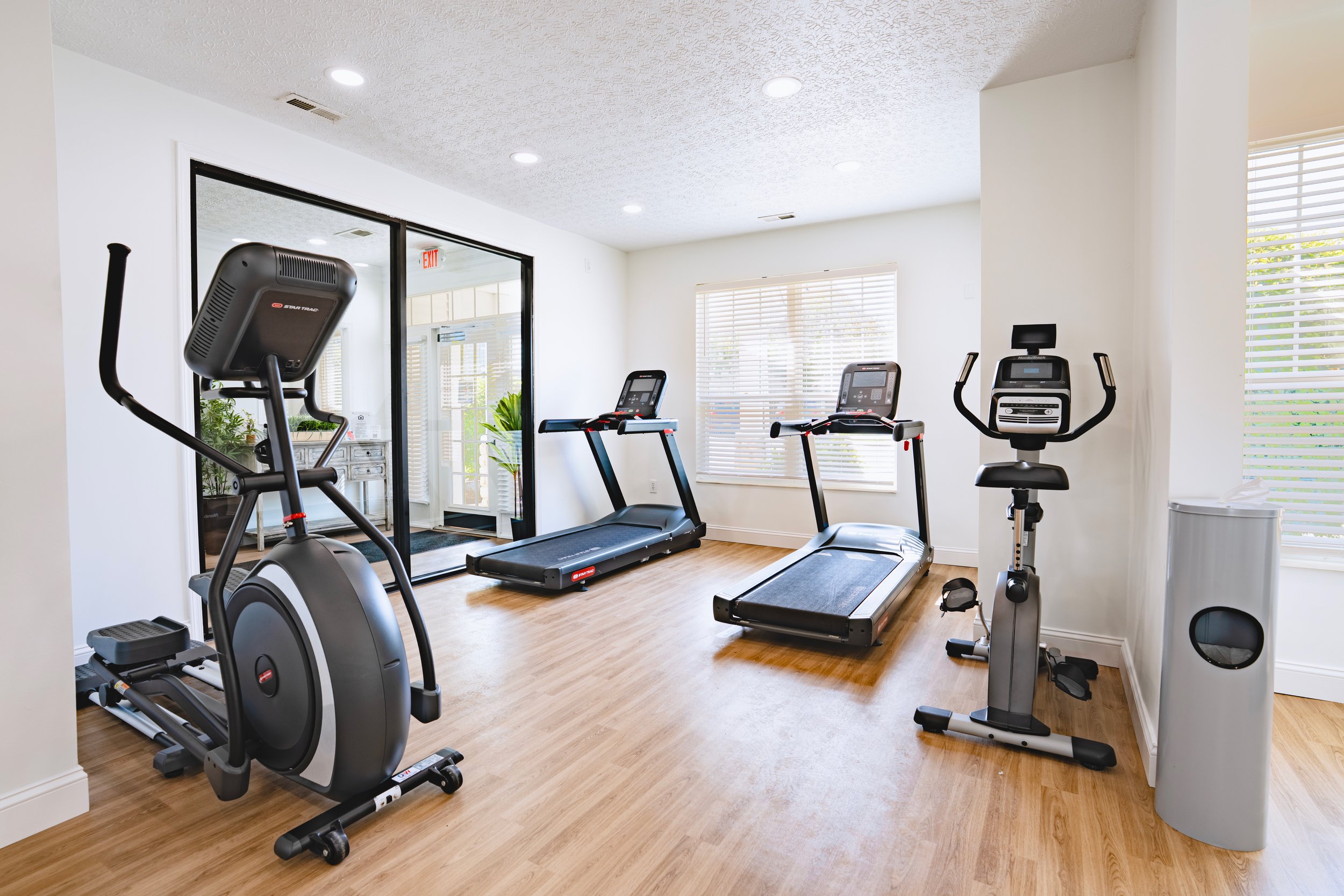 A small home gym with a treadmill, elliptical, and stationary bike near windows with blinds, illuminated by ceiling lights.