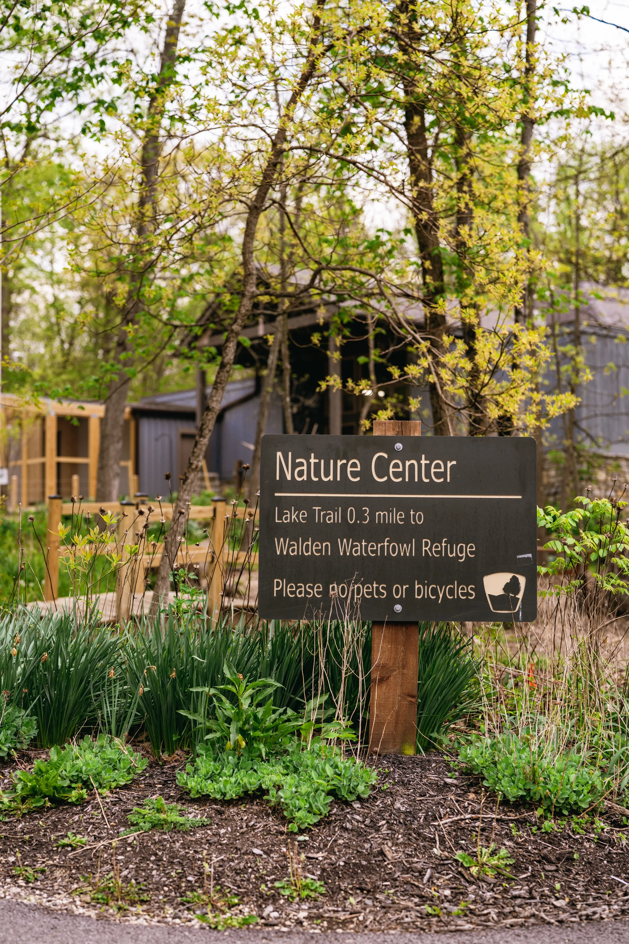 A sign at a nature center with a background of trees and a building, surrounded by green plants and soil.