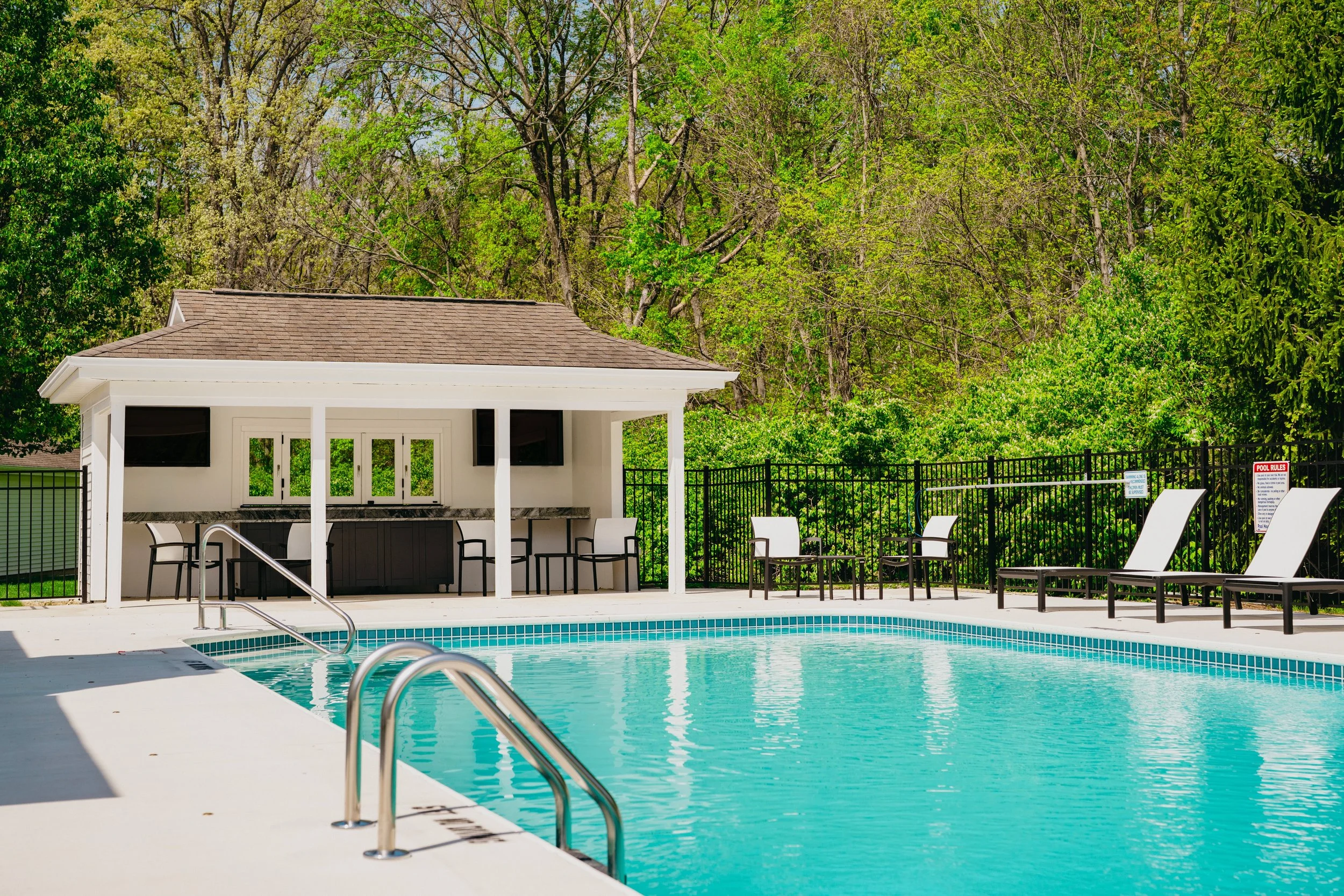 An outdoor swimming pool area with poolside chairs, a poolside shelter with a bar and seating, black fence, and lush green trees in the background.