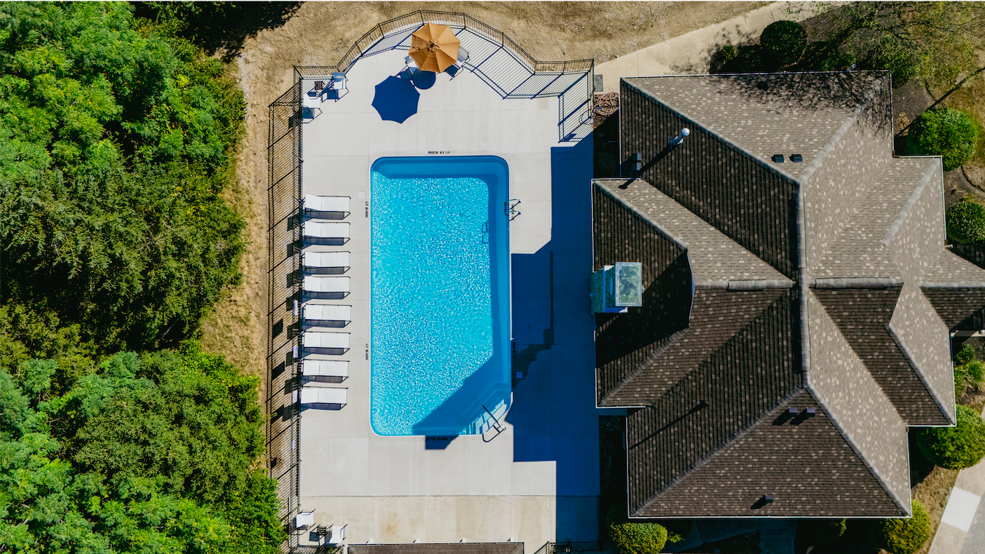 Top-down aerial view of a rectangular swimming pool with white lounge chairs and a beige umbrella on the deck, surrounded by a black metal fence, adjacent to a house with a gray shingled roof, with green trees on the left side.