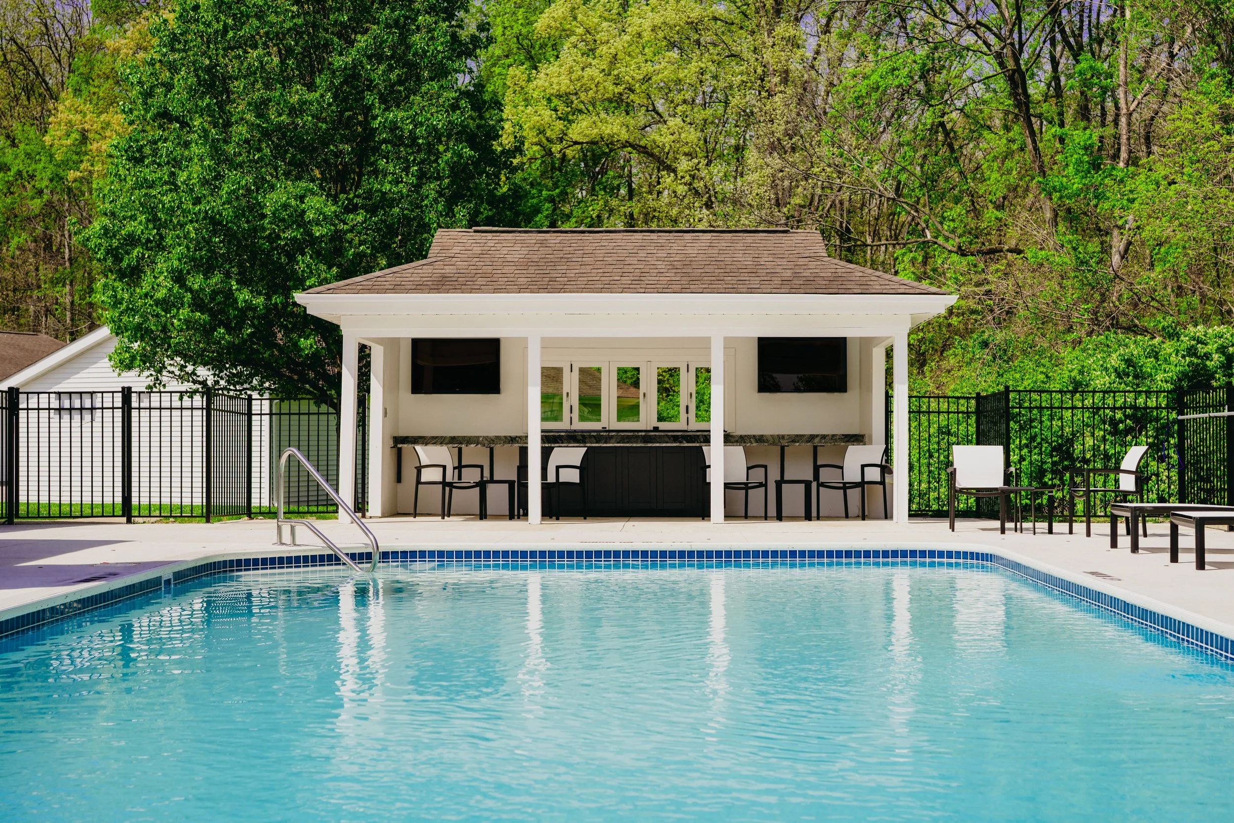 A backyard swimming pool with clear blue water, surrounded by a white concrete deck, with a poolside bar and seating area under a roofed structure, and lush green trees in the background.