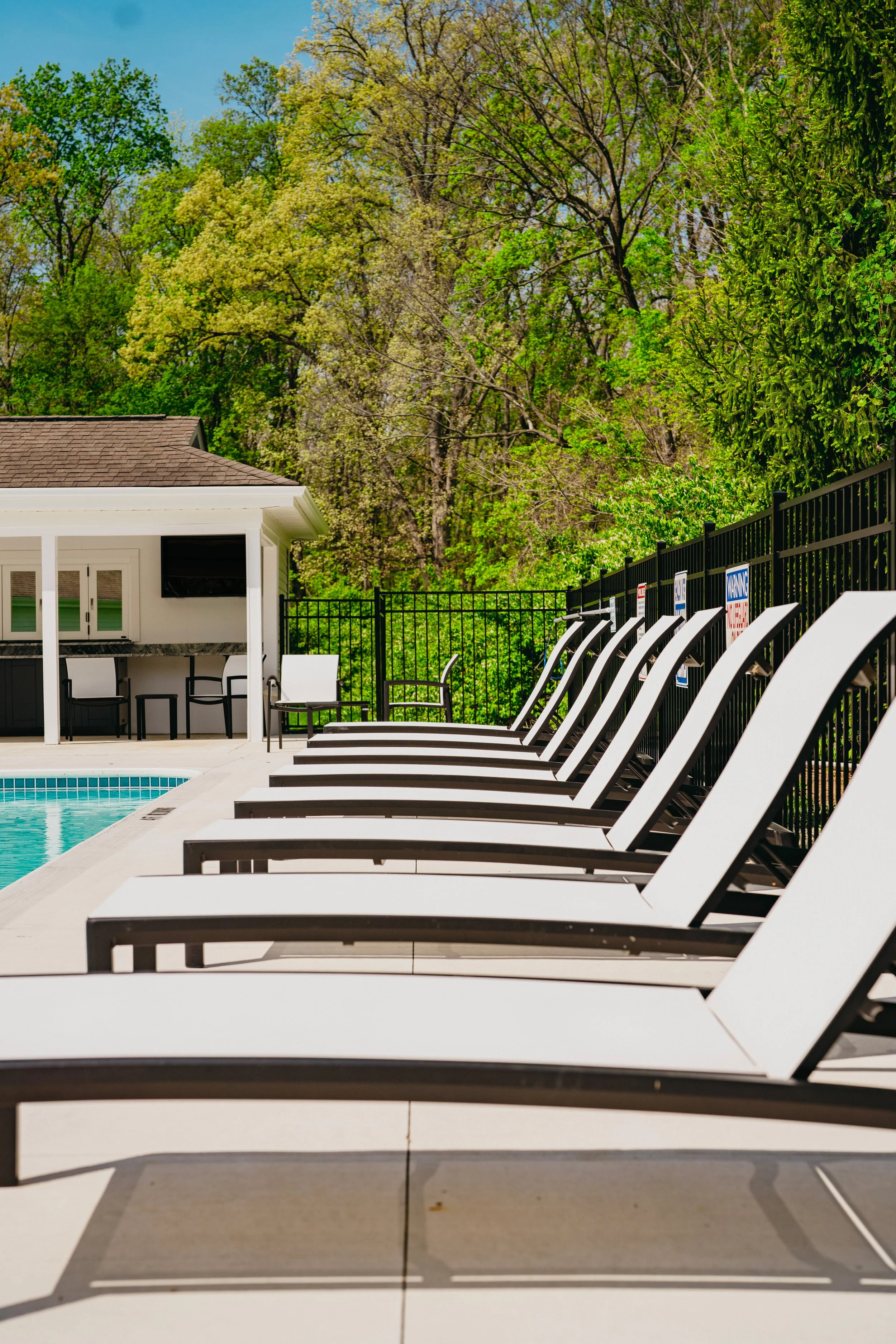 Empty poolside area with lounge chairs, a small building with a bar and televisions, and a view of green trees under a blue sky.