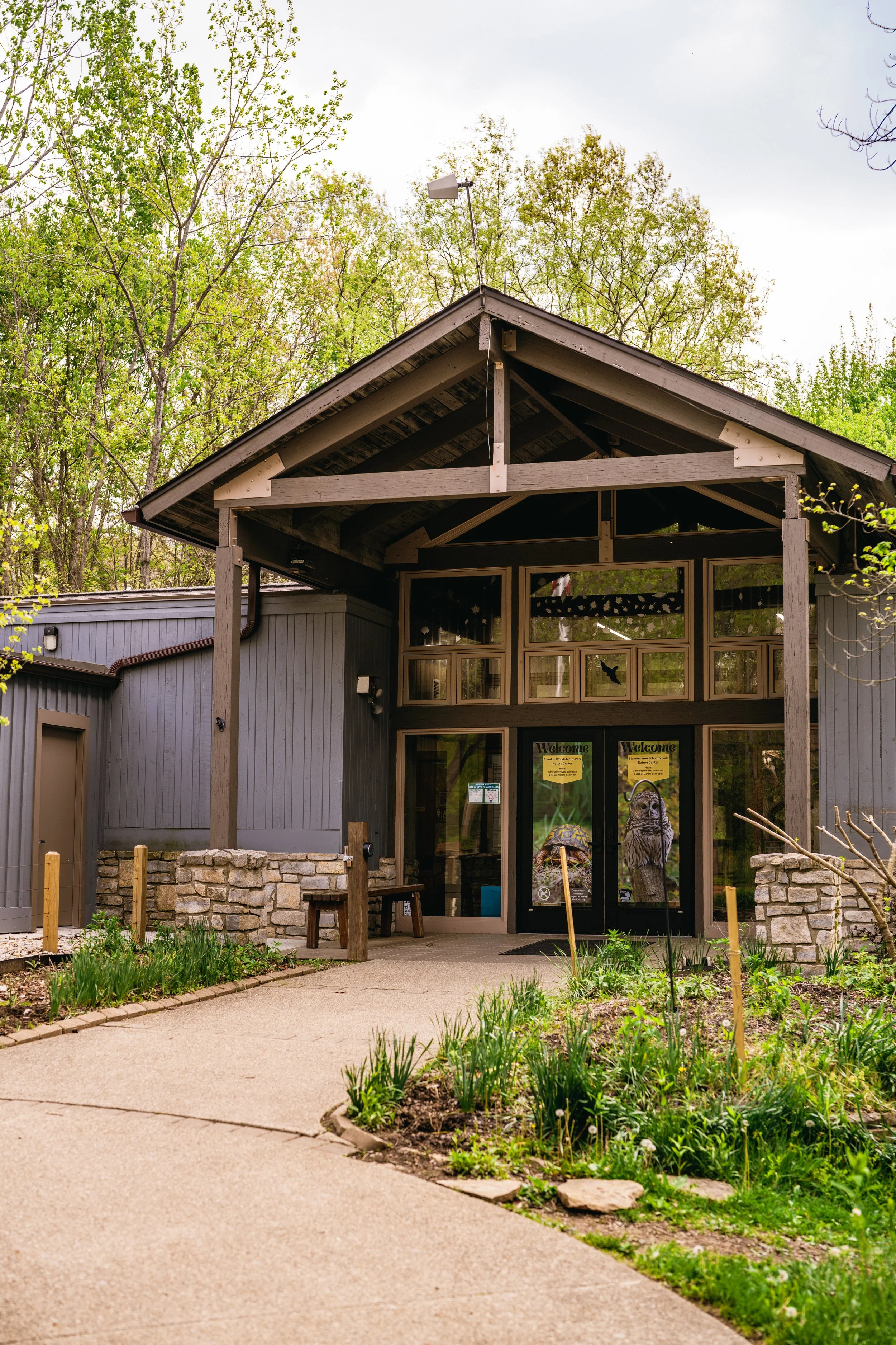 Entrance of a building with stone and wooden exterior, surrounded by a garden with young plants, and trees in the background.