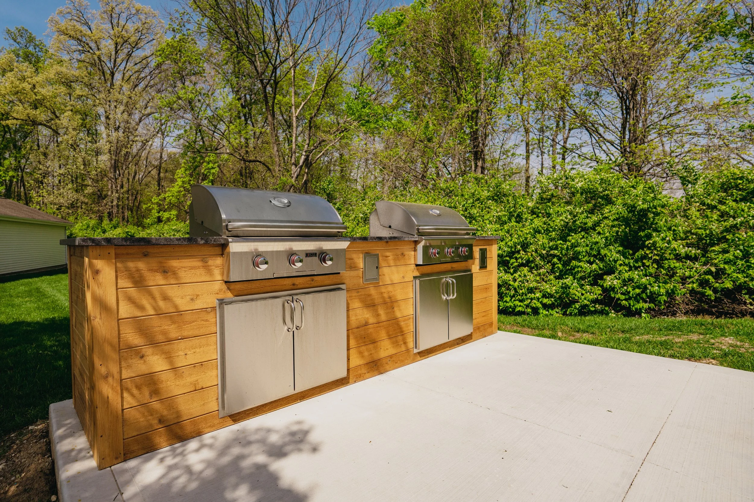 Outdoor kitchen with stainless steel grills and wooden cabinetry on a concrete slab, surrounded by green trees and a grassy lawn on a clear, sunny day.