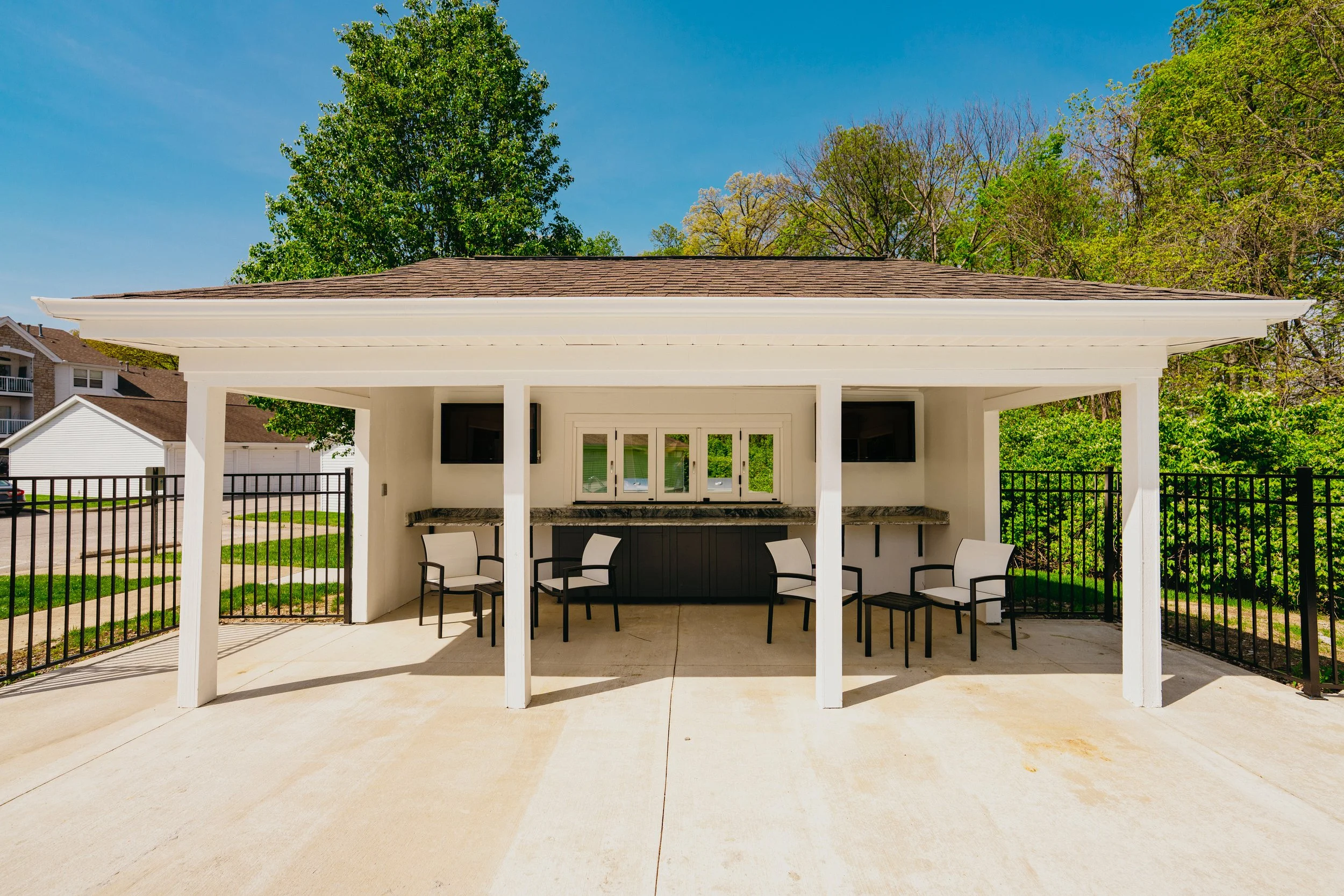 A white outdoor pavilion with a brown shingled roof, surrounded by a black metal fence, with chairs and a counter inside, and trees in the background under a clear blue sky.