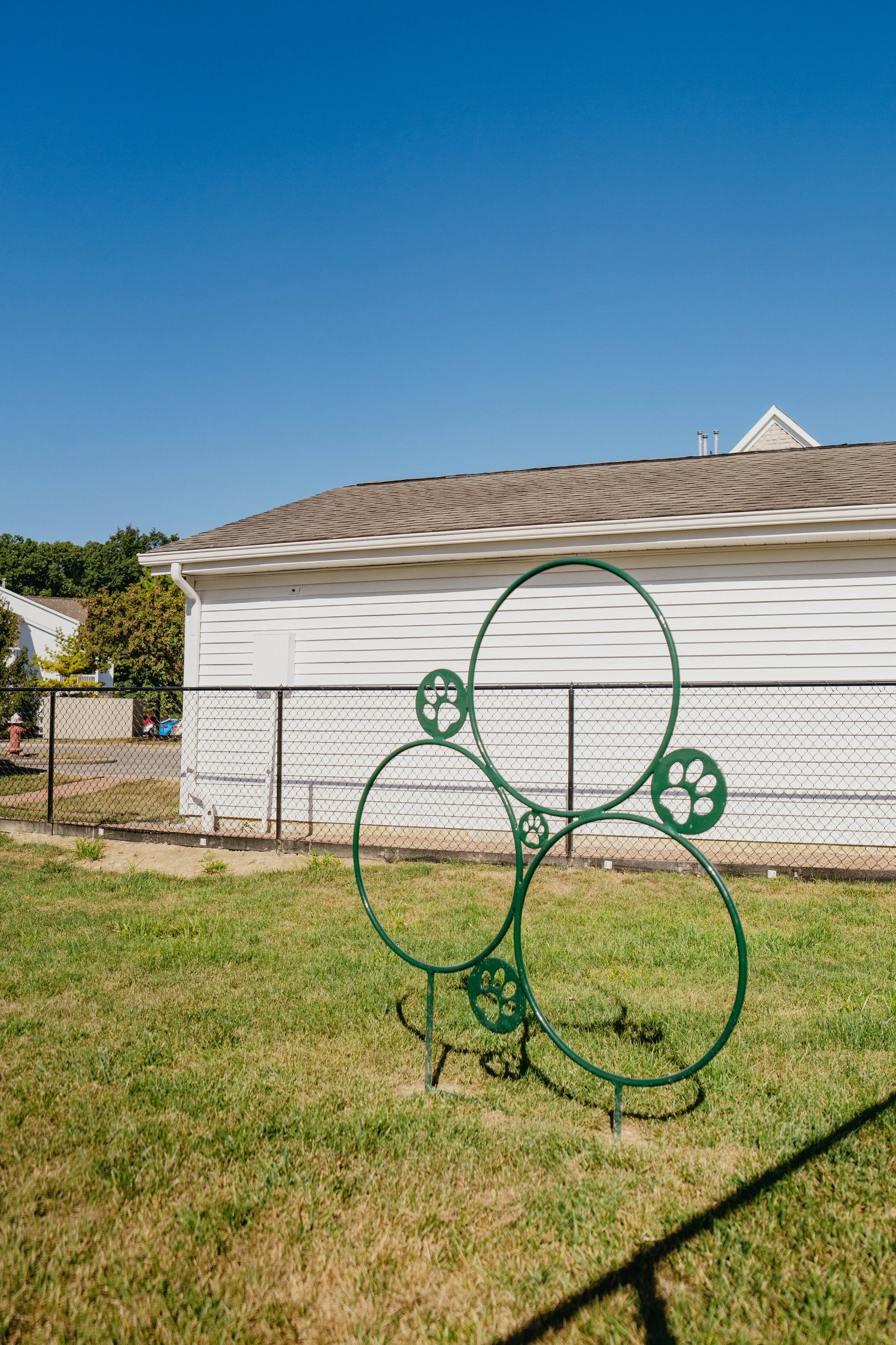 A playground with a green metal climbing structure in the shape of interconnected loops and circles. A white house with beige roof and gutter is in the background, with a clear blue sky overhead. The playground is enclosed by a chain-link fence, and 