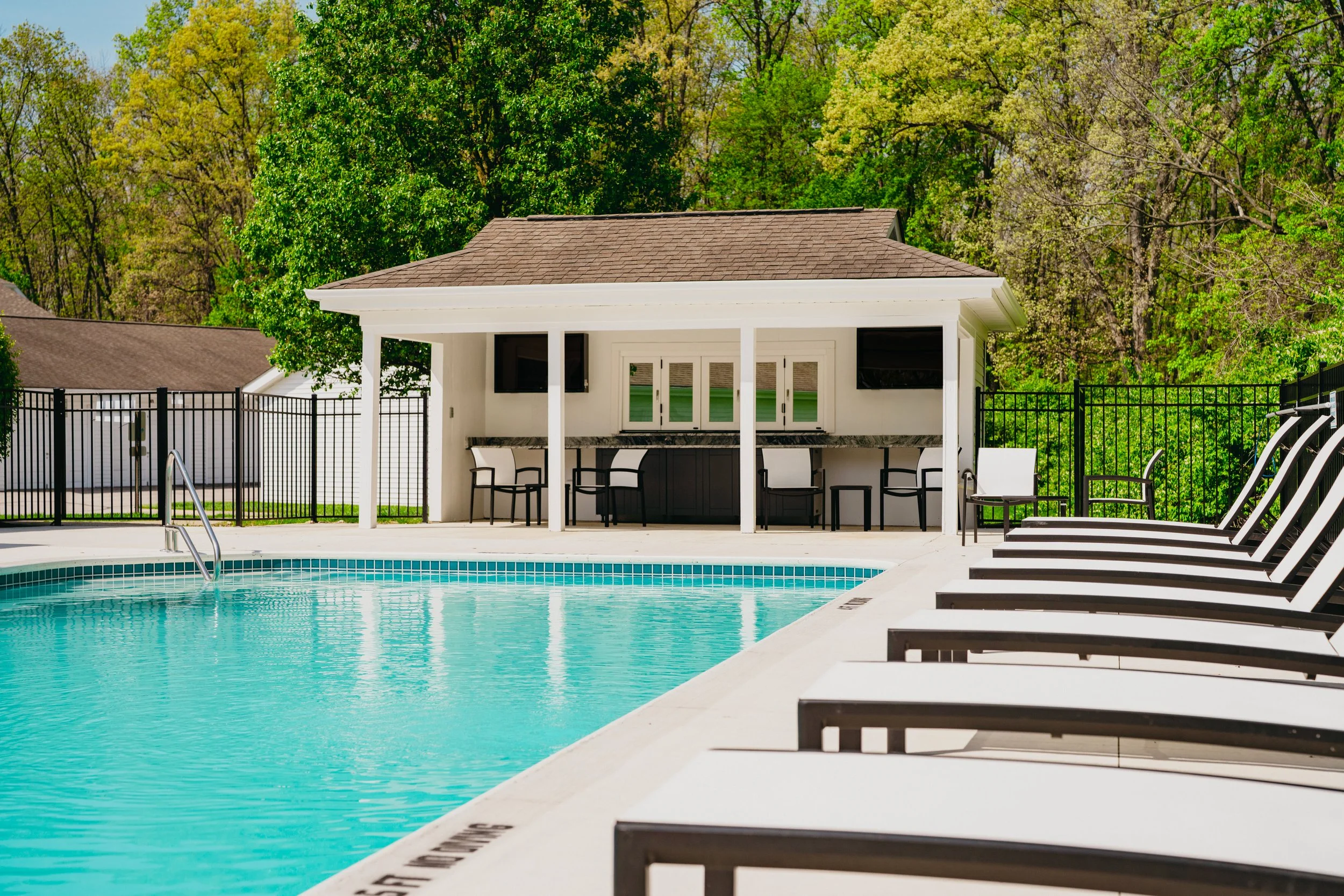 Empty swimming pool with lounge chairs along the edge, and a pool house with outdoor bar area, surrounded by trees.