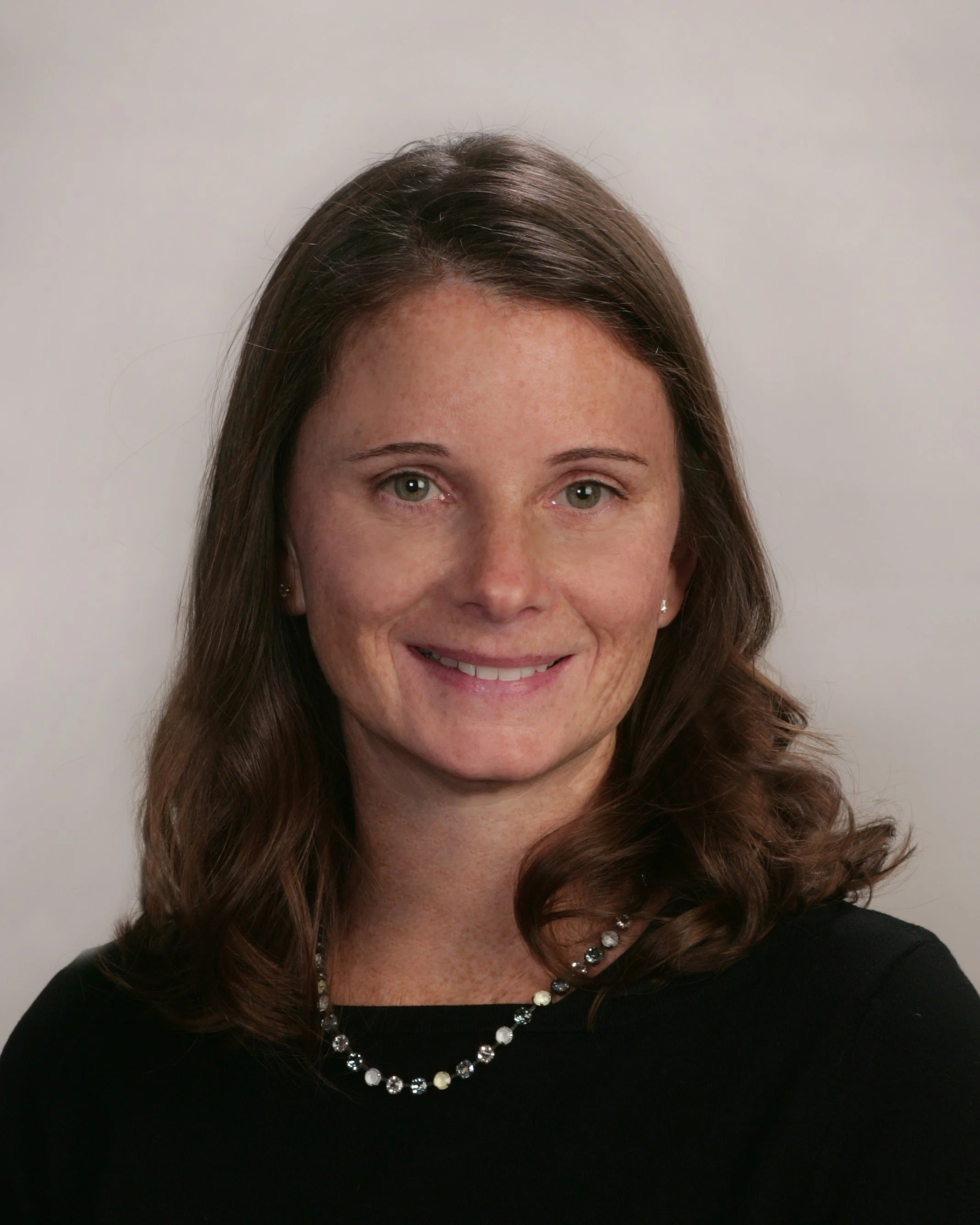 Headshot of a woman with shoulder-length brown hair, wearing a black top and a pearl necklace, smiling against a plain light-colored background.