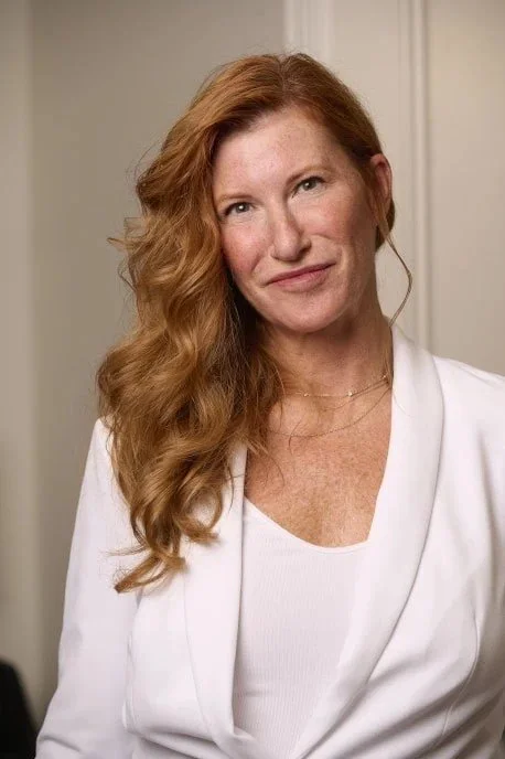 A woman with long wavy red hair, wearing a white blazer and a white top, standing indoors near a door.