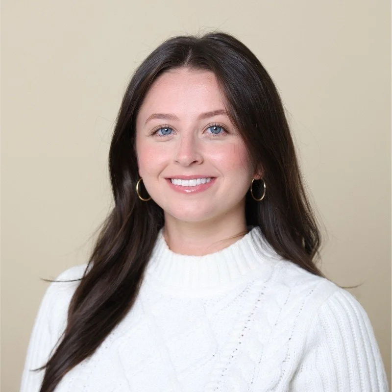 A portrait of a young woman with long dark brown hair, blue eyes, wearing gold hoop earrings and a white knit sweater, smiling against a beige background.