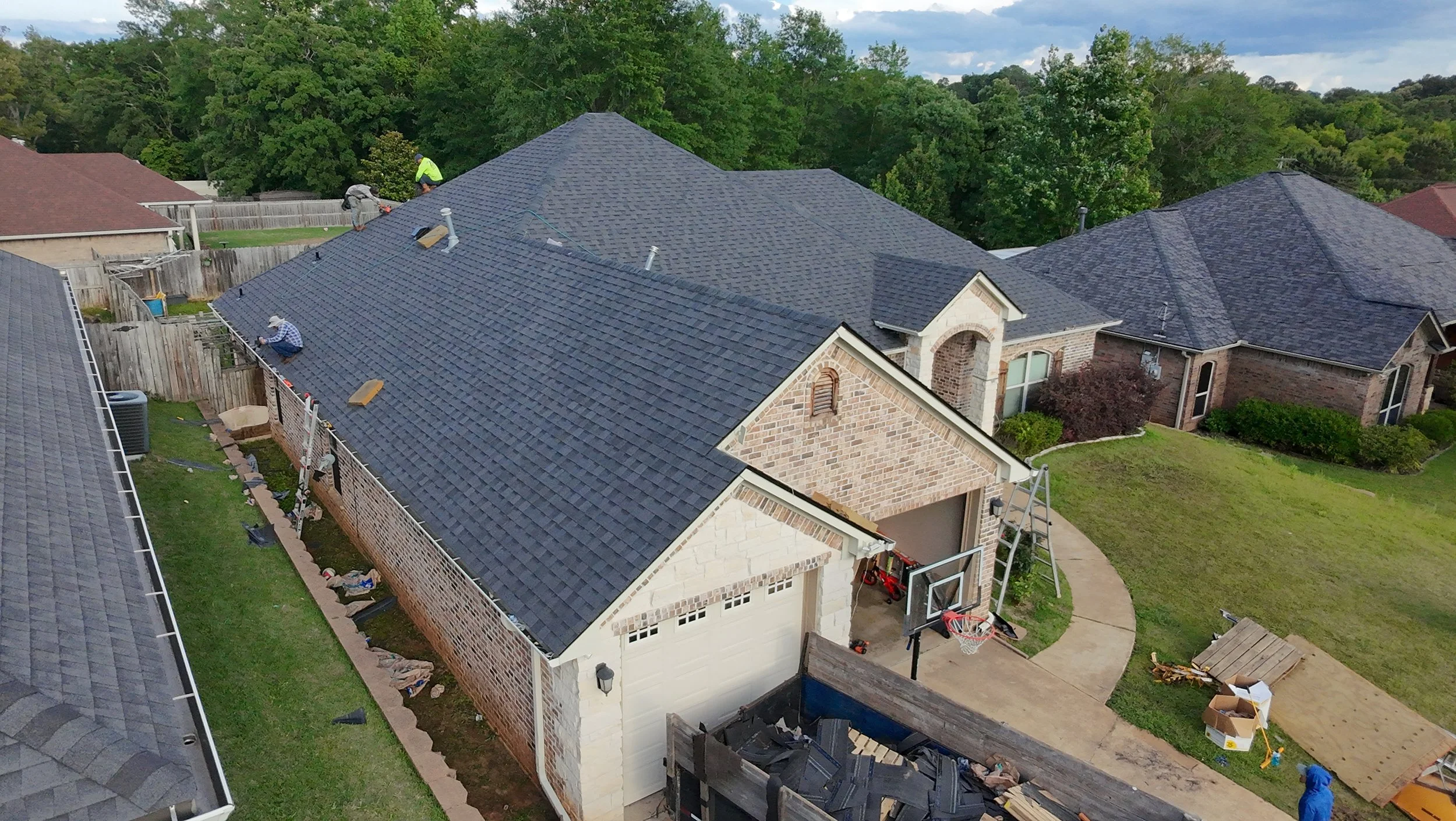 Aerial view of a house with people working on the roof, replacing or repairing shingles, with a backyard, basketball hoop, and lawn.