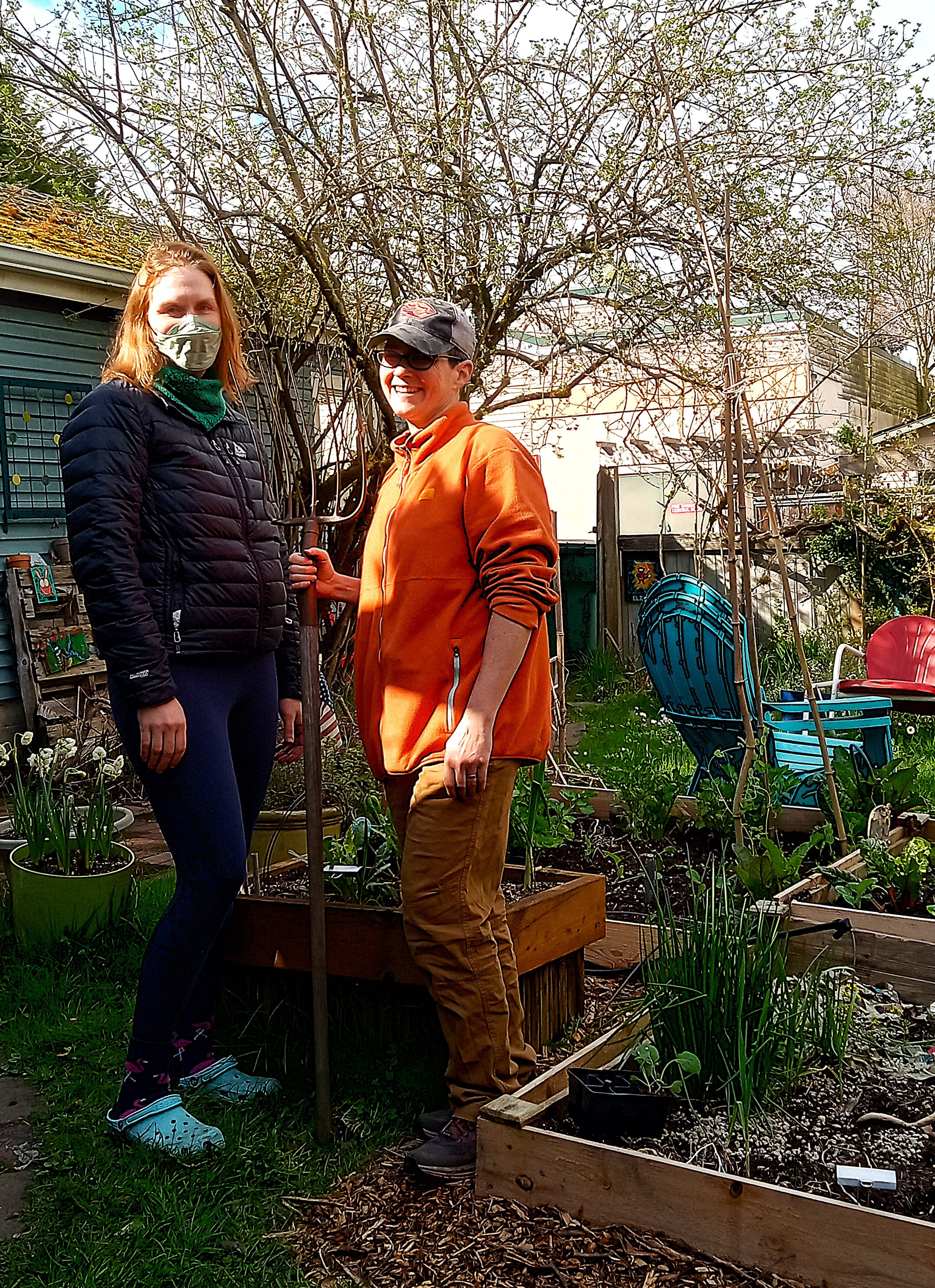 Two women posing in a backyard with raised beds and patio chairs in the background. One holds a pitchfork.