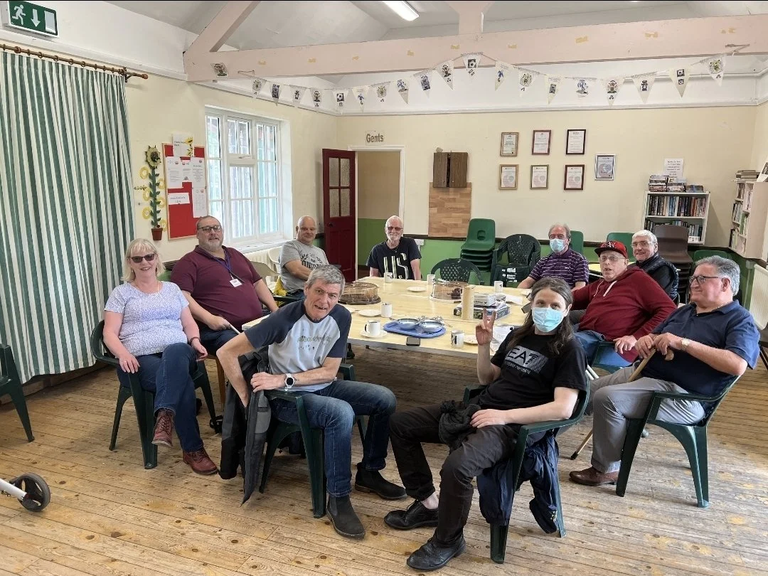 A group of eleven adults sitting around a rectangular table in a community room, some wearing masks, with cakes, mugs, and plates on the table, and a few plants and certificates on the walls.