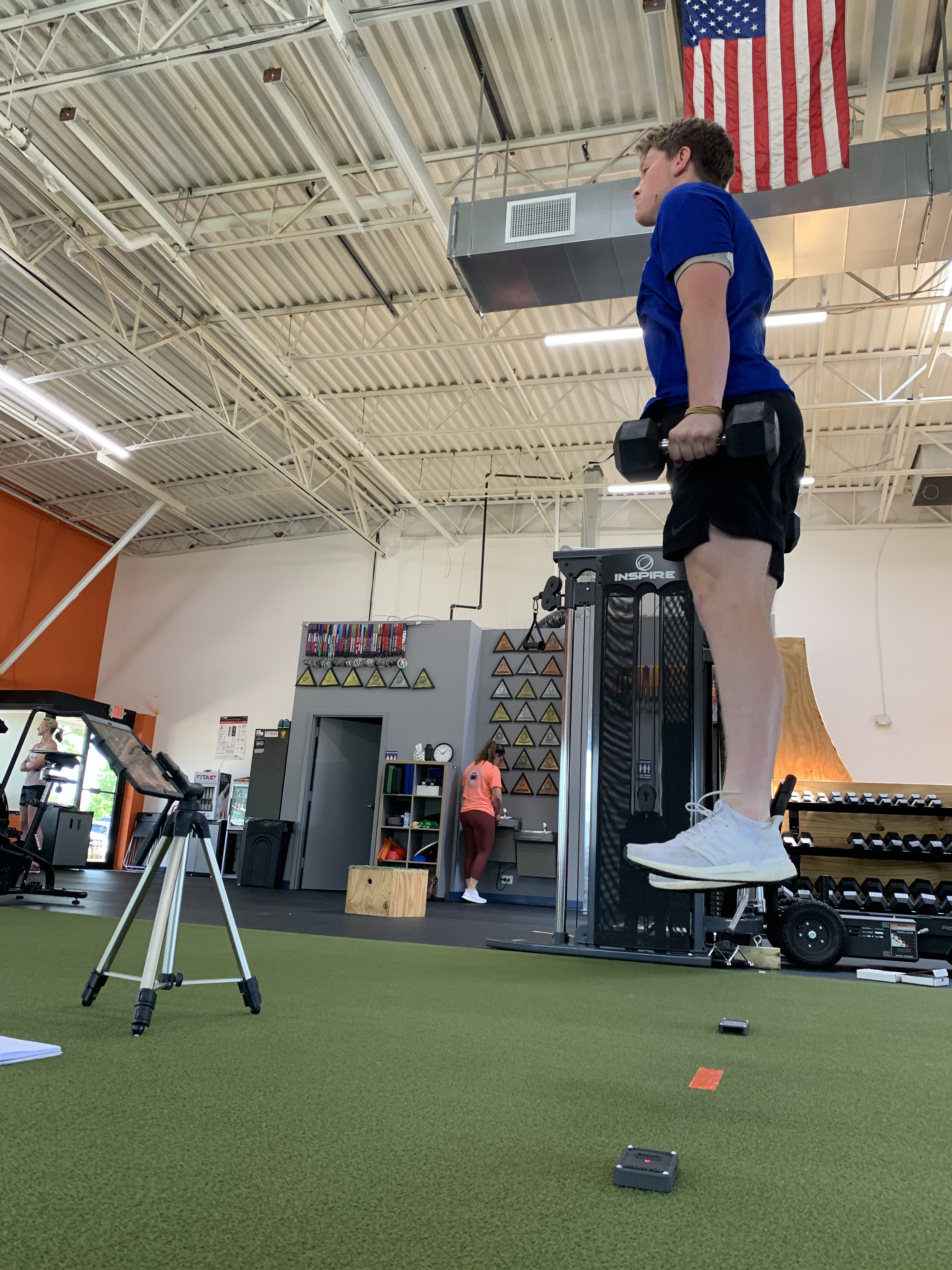 A person in a gym performing a jump exercise with dumbbells, under an American flag, with gym equipment in the background.
