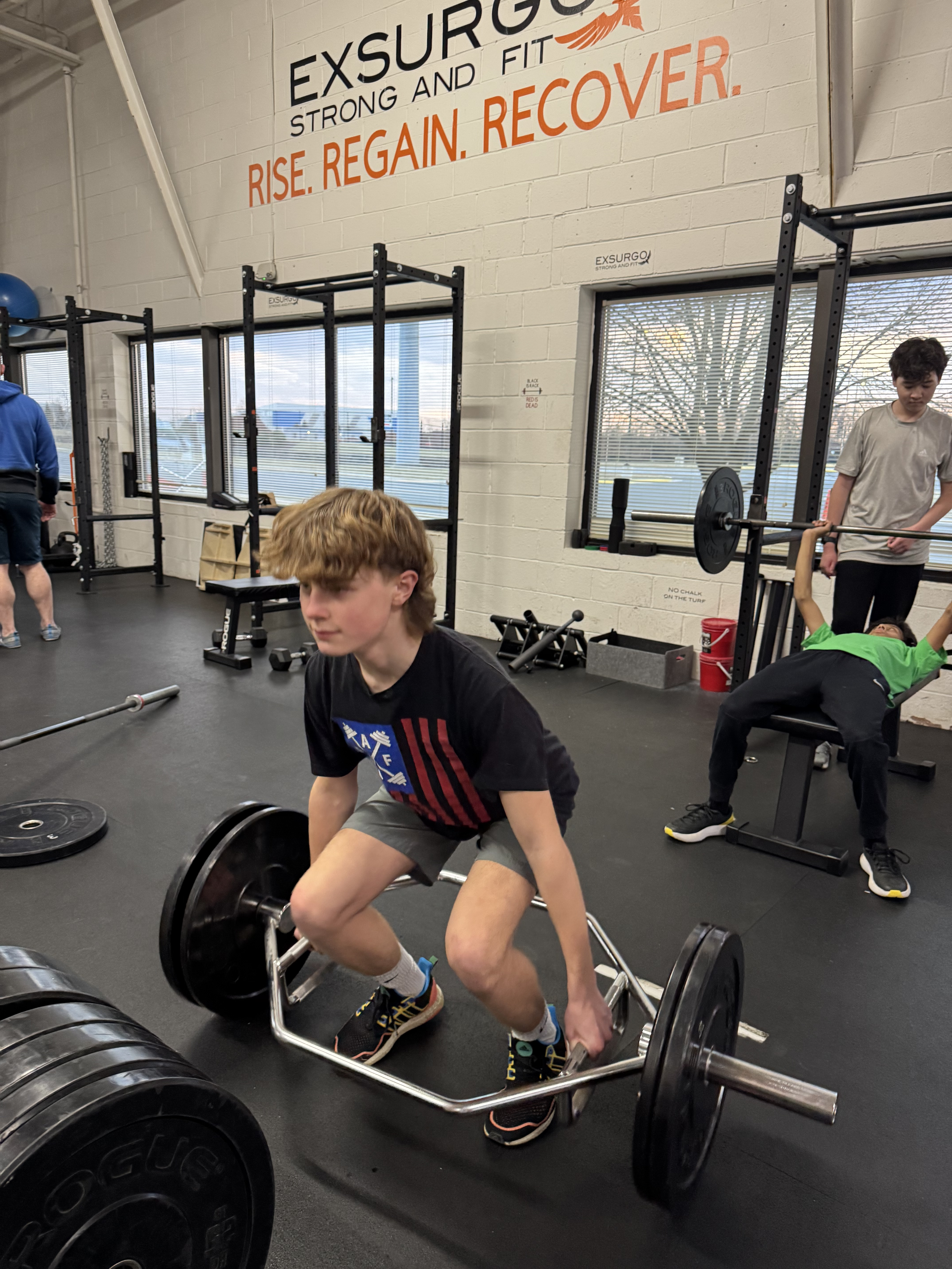 Person lifting weights with a trap bar at a gym, another person bench pressing in the background.
