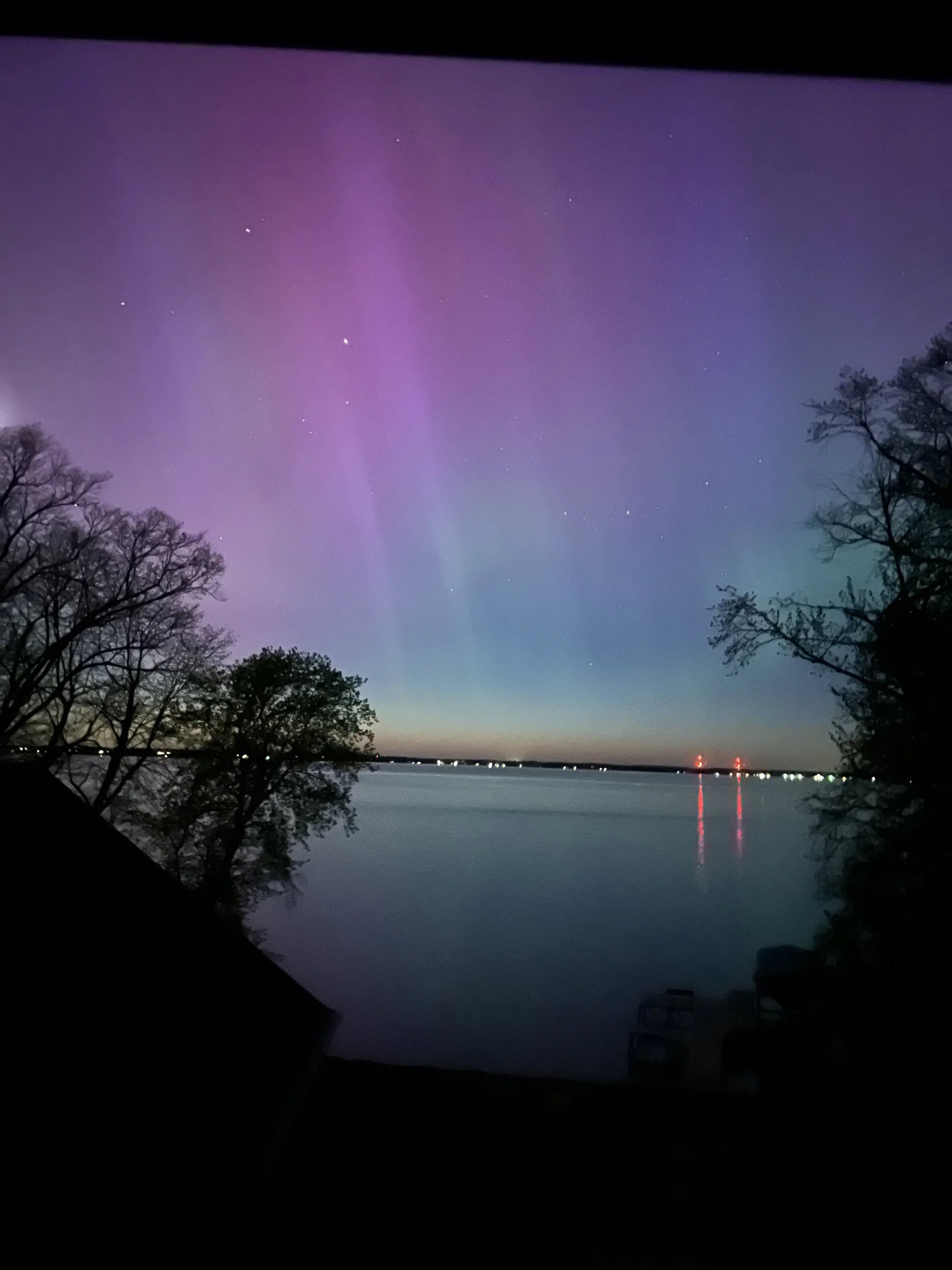 Aurora borealis over a calm lake with silhouetted trees and distant city lights.