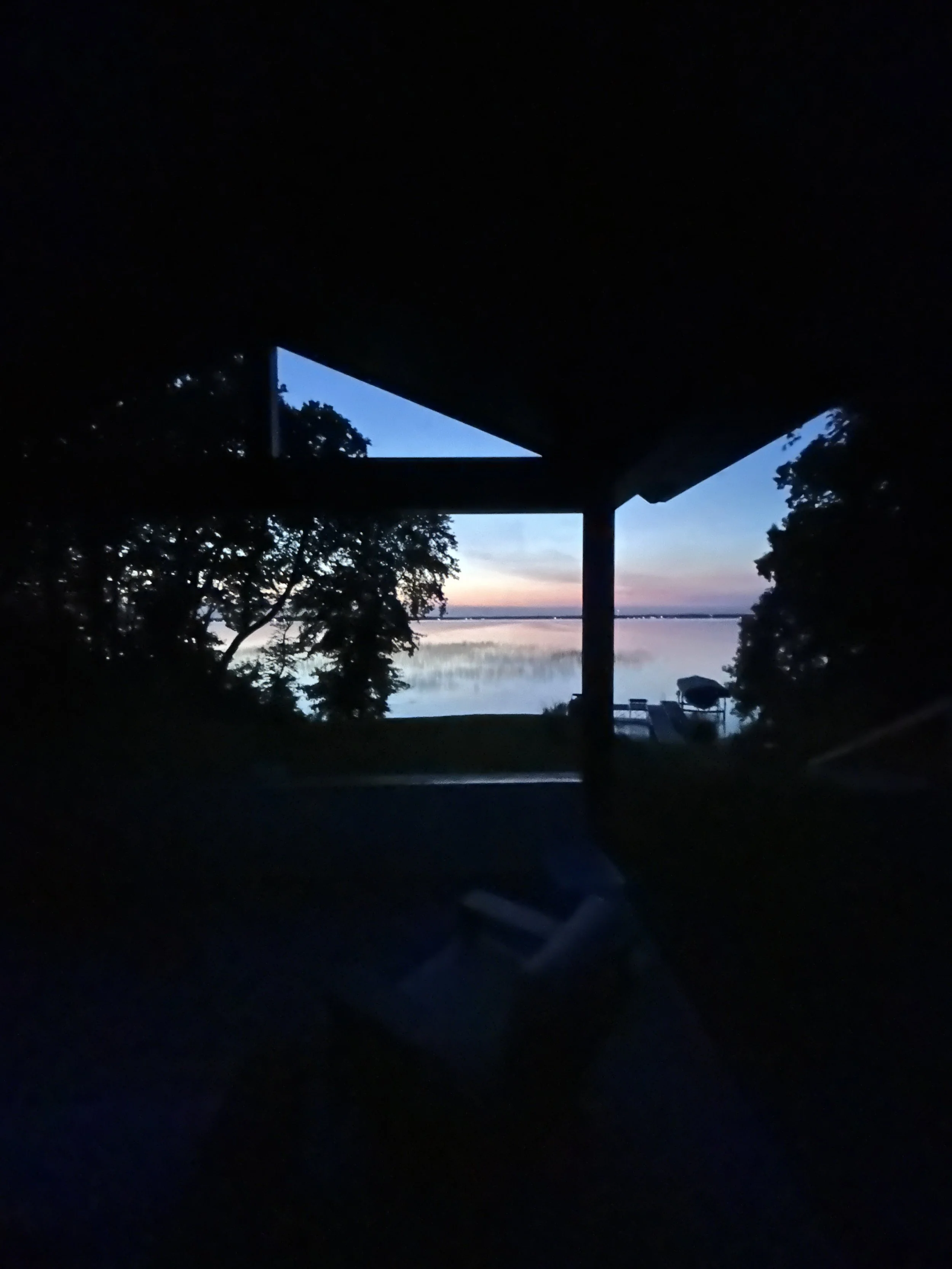 Silhouette of trees and lake visible through an open window at sunset, dark interior.