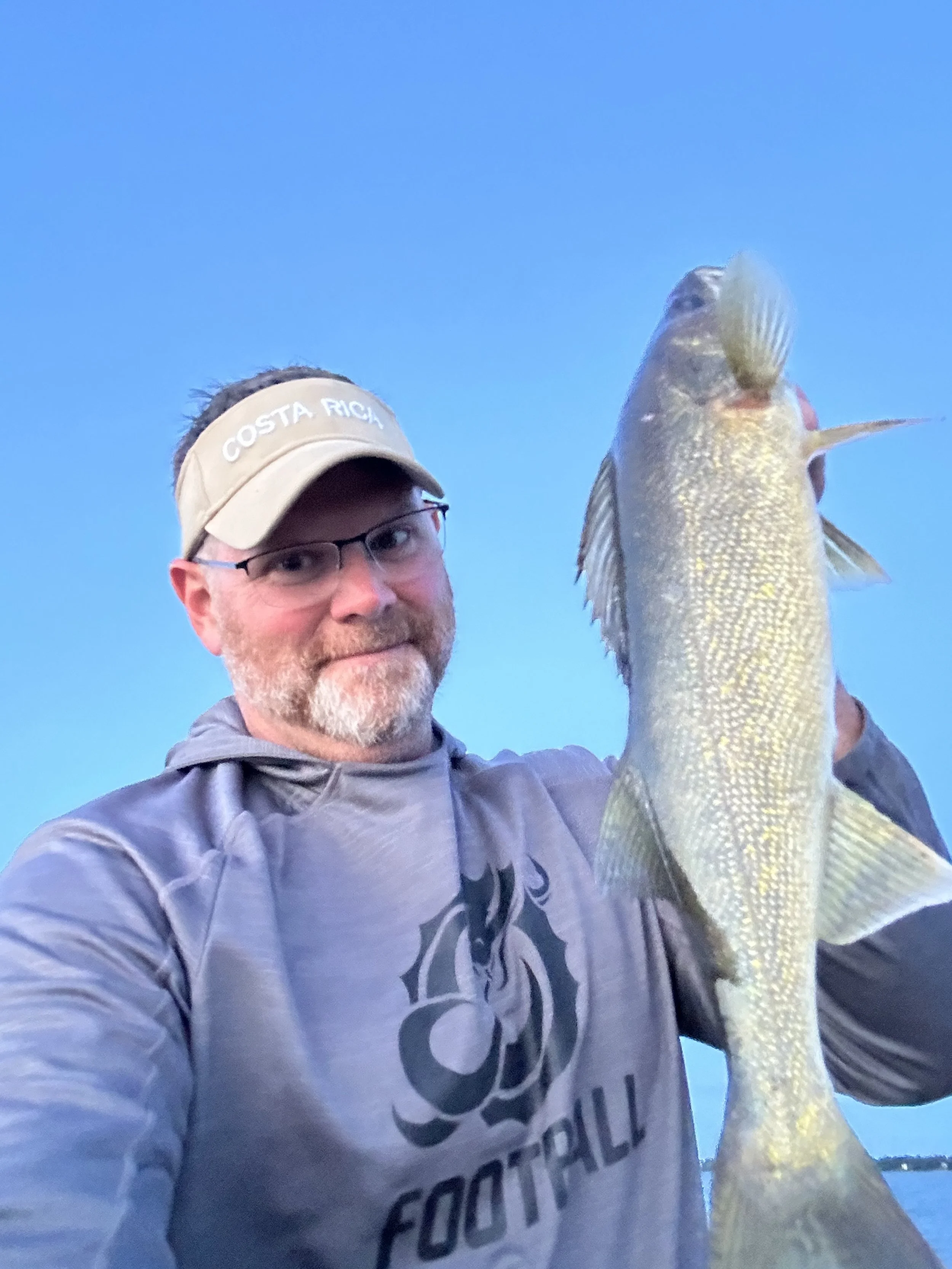 Man holding a fish outdoors, wearing a visor and glasses, with a blue sky background.