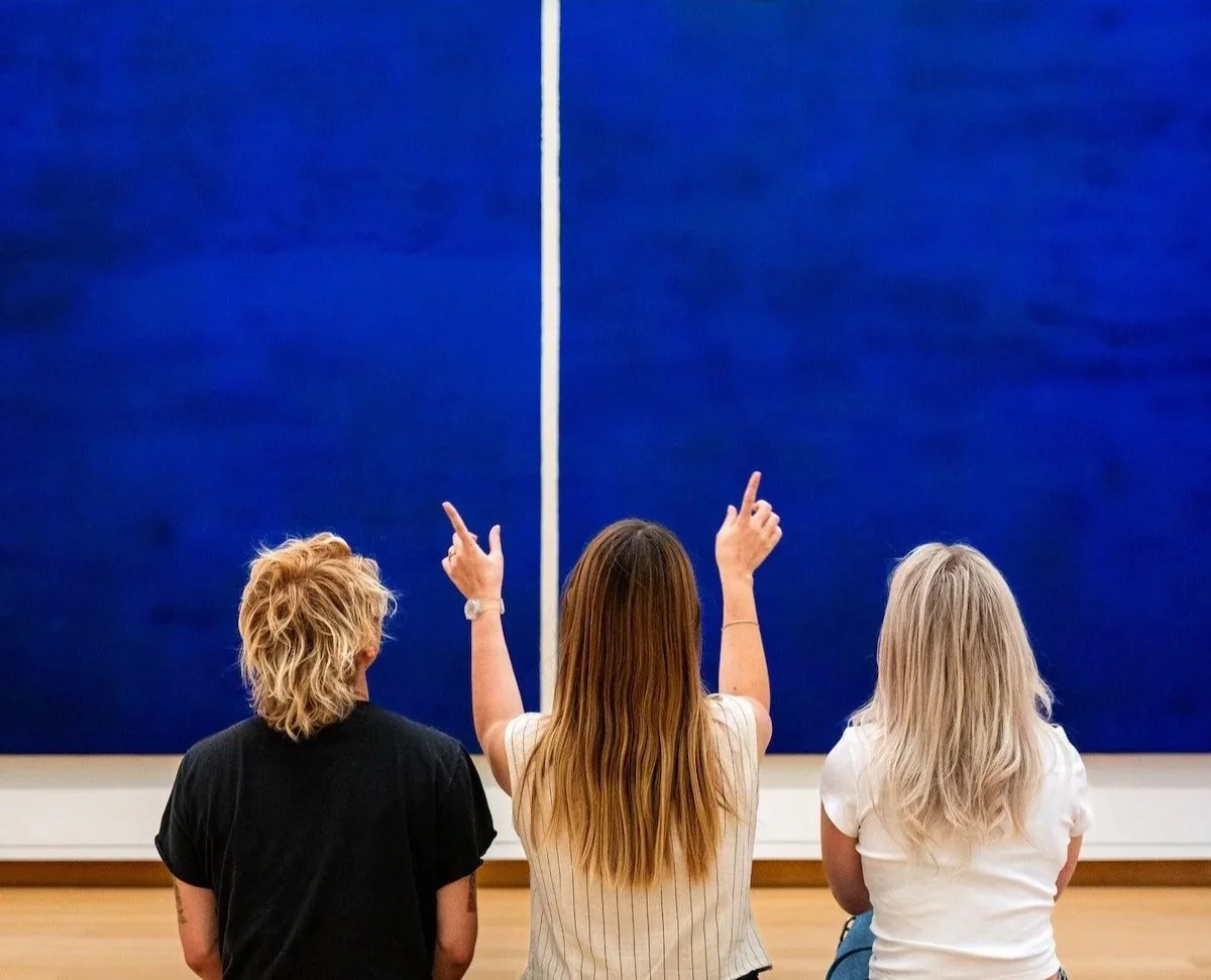 Three women with different hair colors and styles, sitting and observing a large blue art piece in a gallery. The woman in the middle is pointing at the artwork.