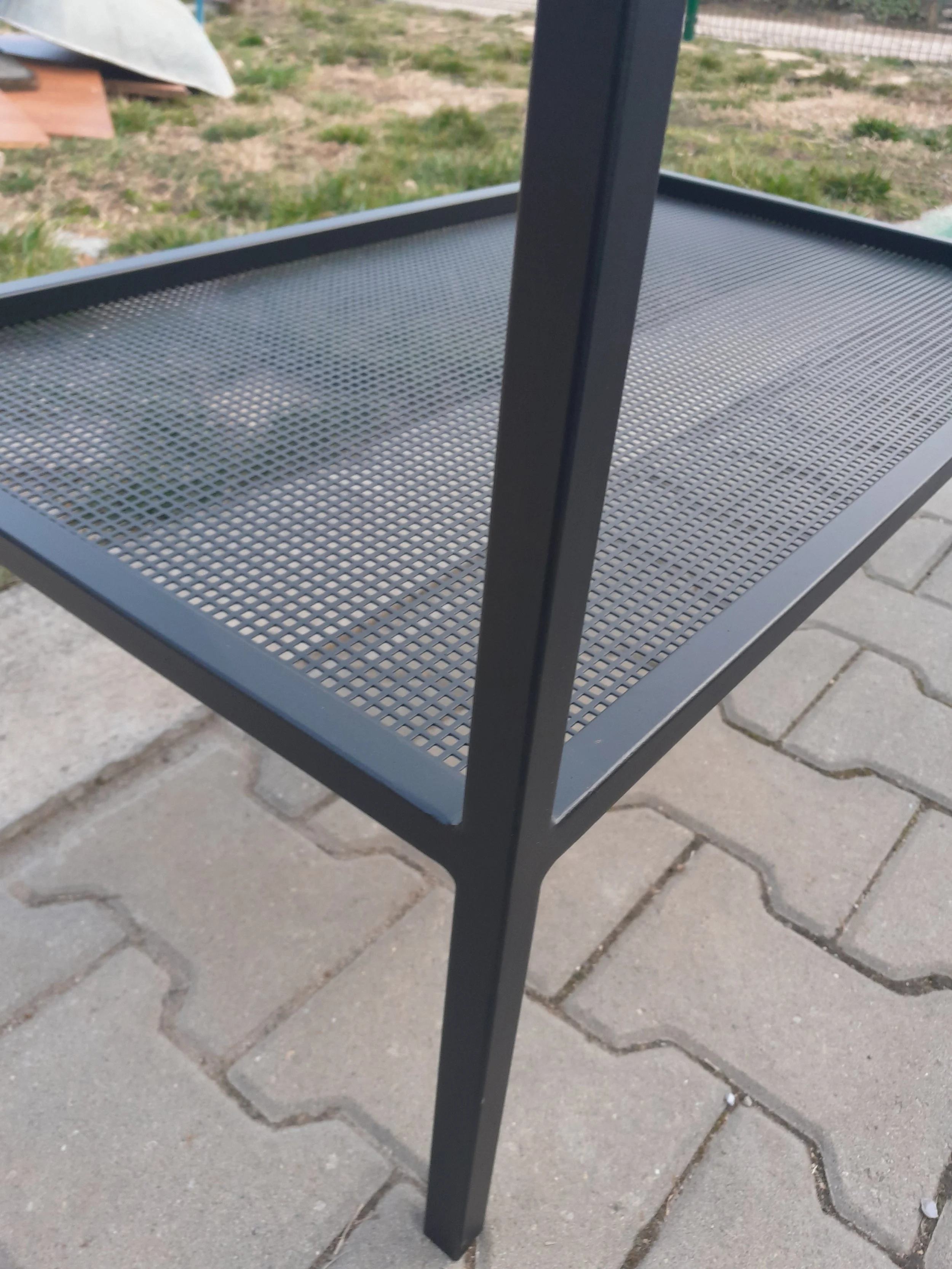 Close-up of a black metal table with perforated mesh shelves, outdoors on a stone patio.