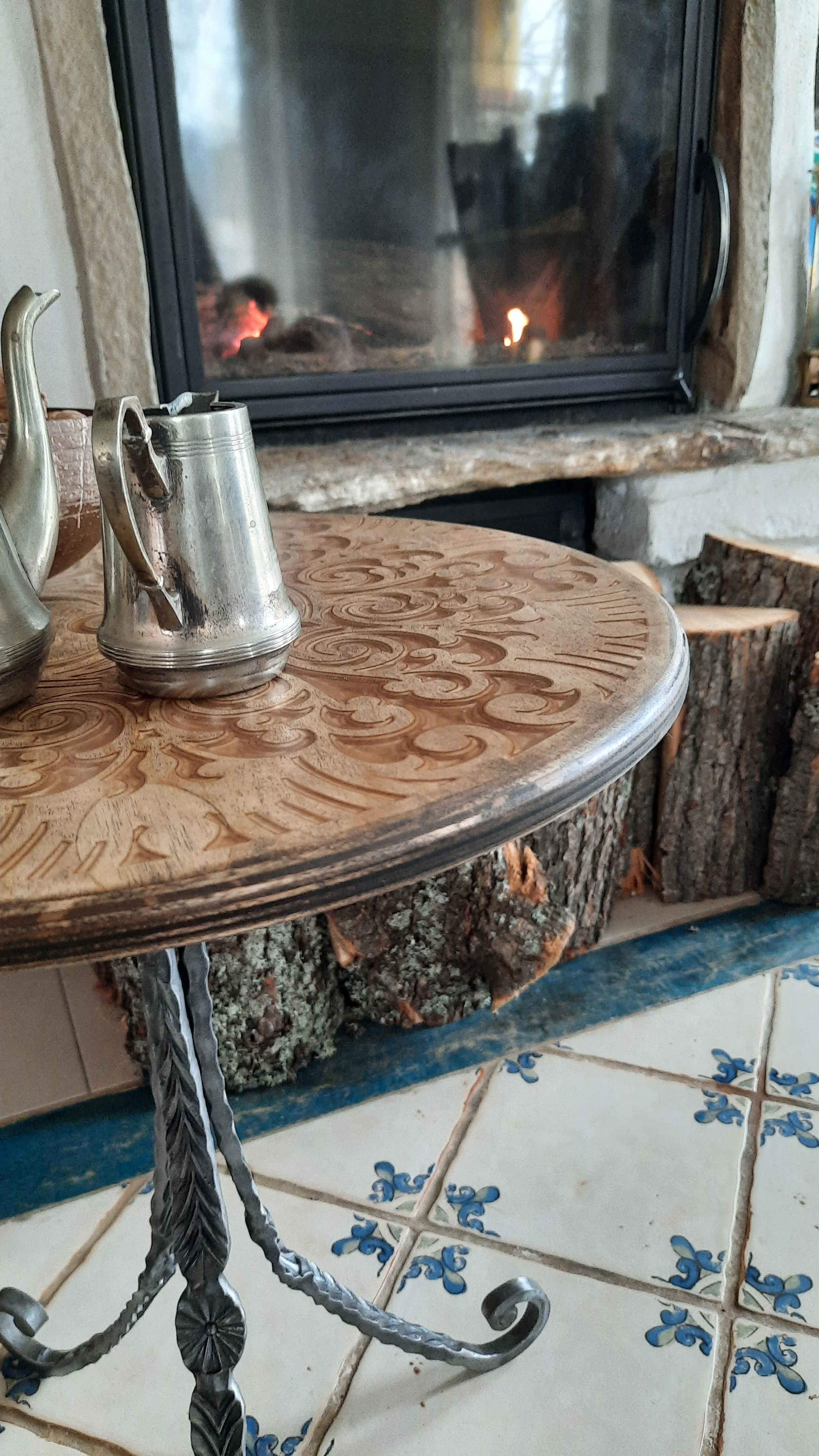 Ornate wooden table with metal kettles near a fireplace and stacked firewood, with tiled floor.