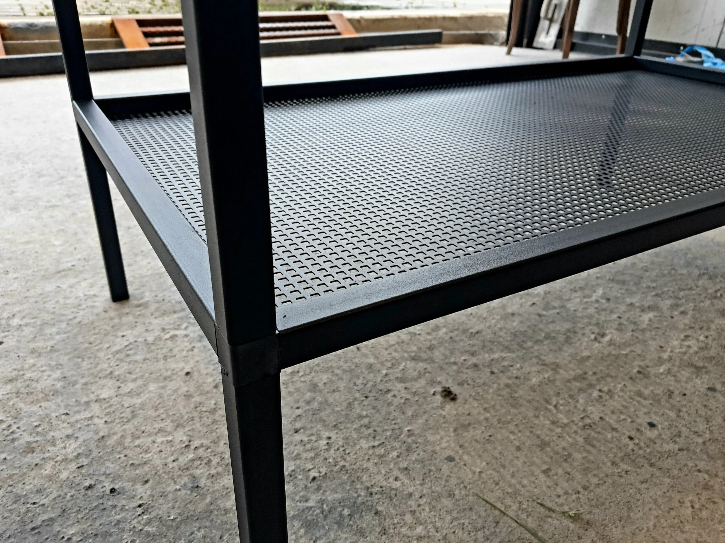 Close-up of a black metal table with a perforated shelf on a concrete floor.