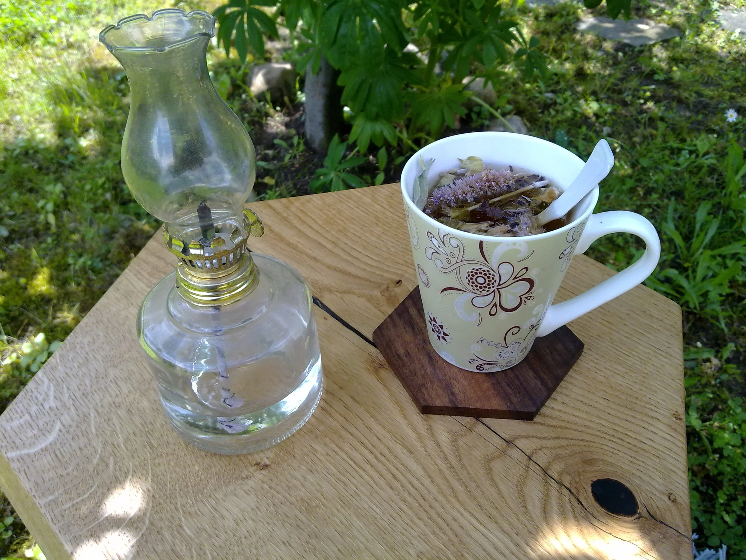 A vintage glass oil lamp next to a floral-patterned mug filled with herbal tea and a spoon on a wooden table outdoors.