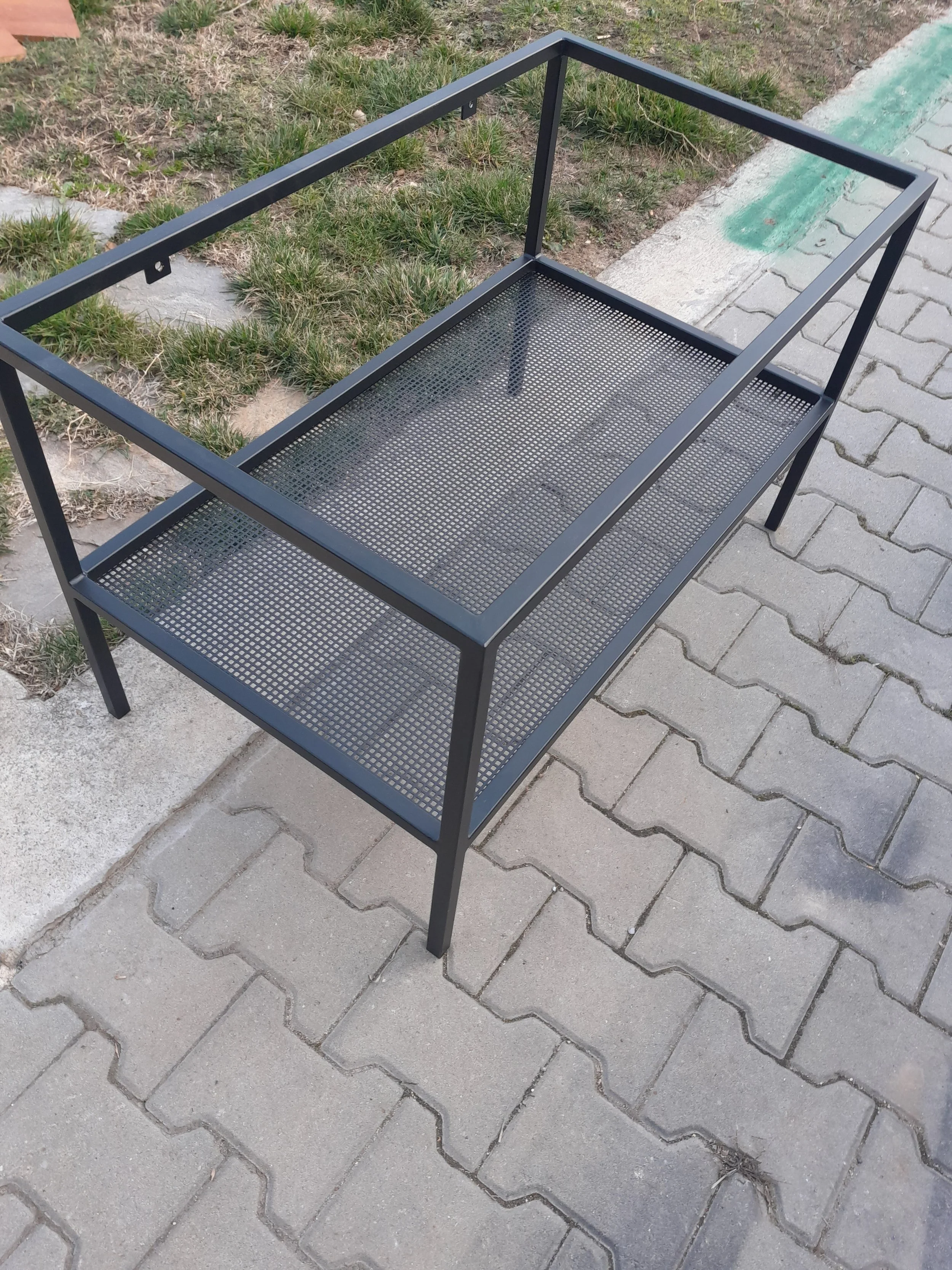 Black metal table frame with mesh shelf on paved and grassy area outdoors