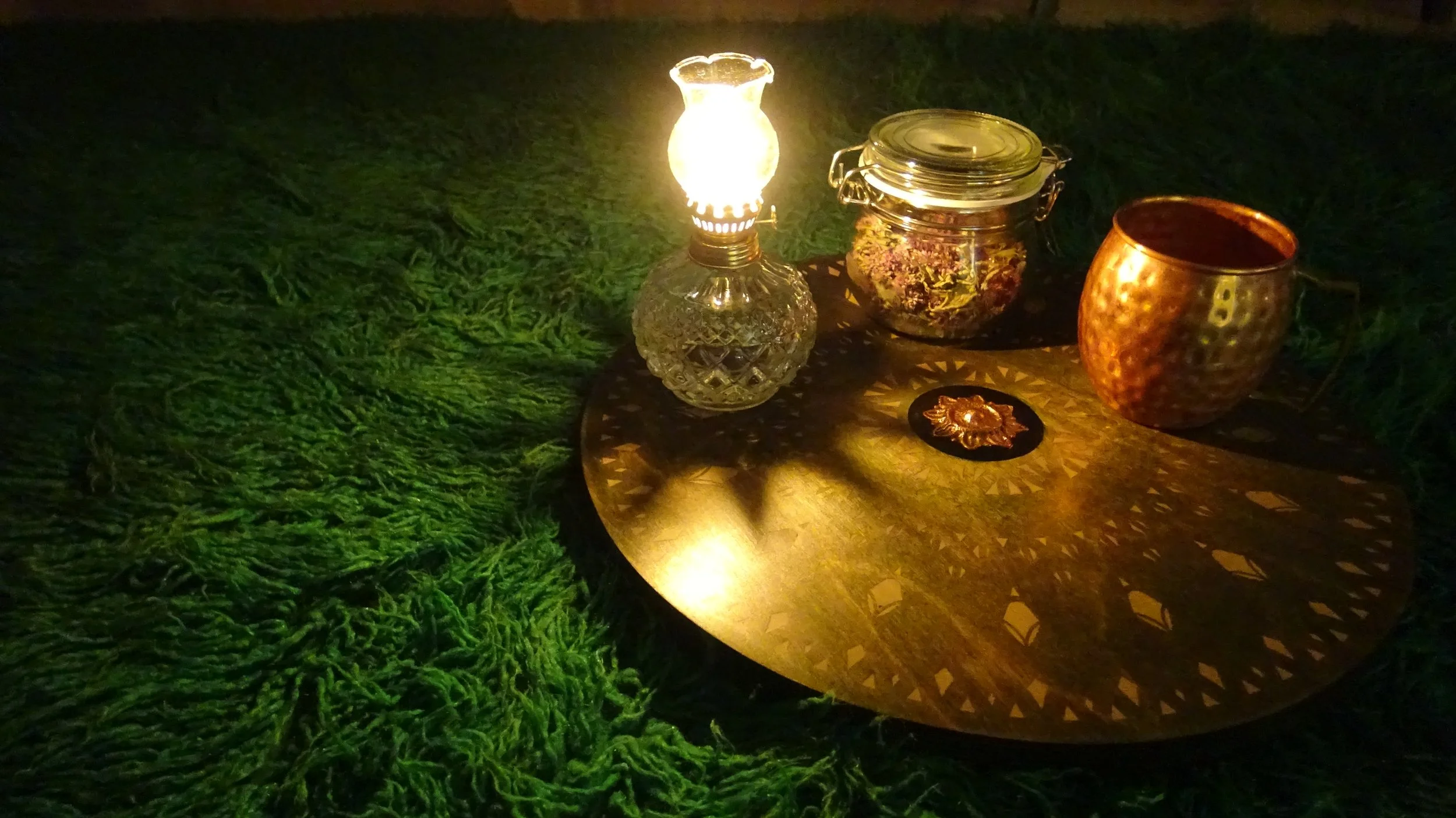 A cozy setting with a lit glass oil lamp, a jar of dried flowers, and a copper mug on a round wooden table, placed on a green textured rug.