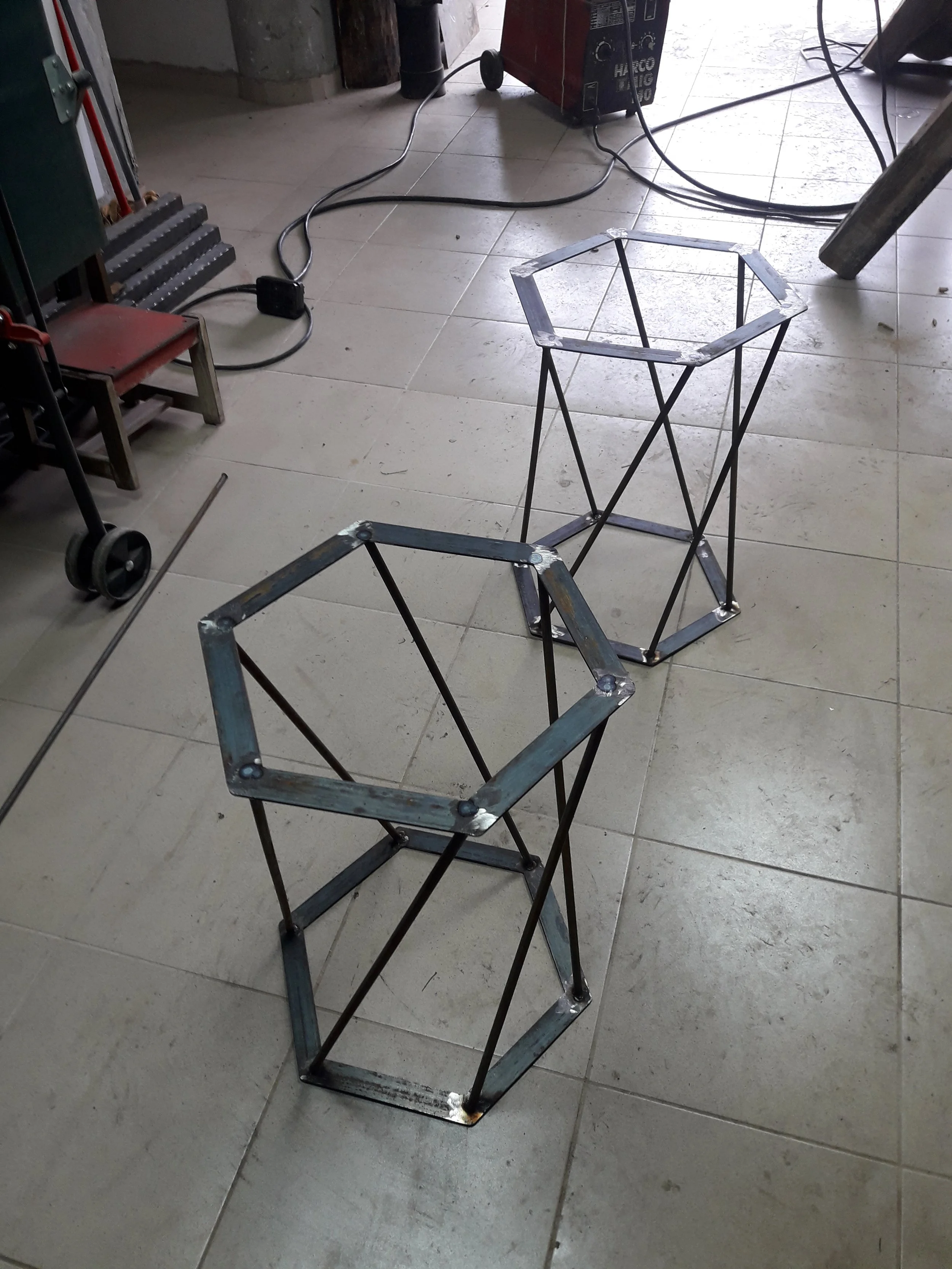 Two hexagonal metal stands on a tiled workshop floor with cables in the background.