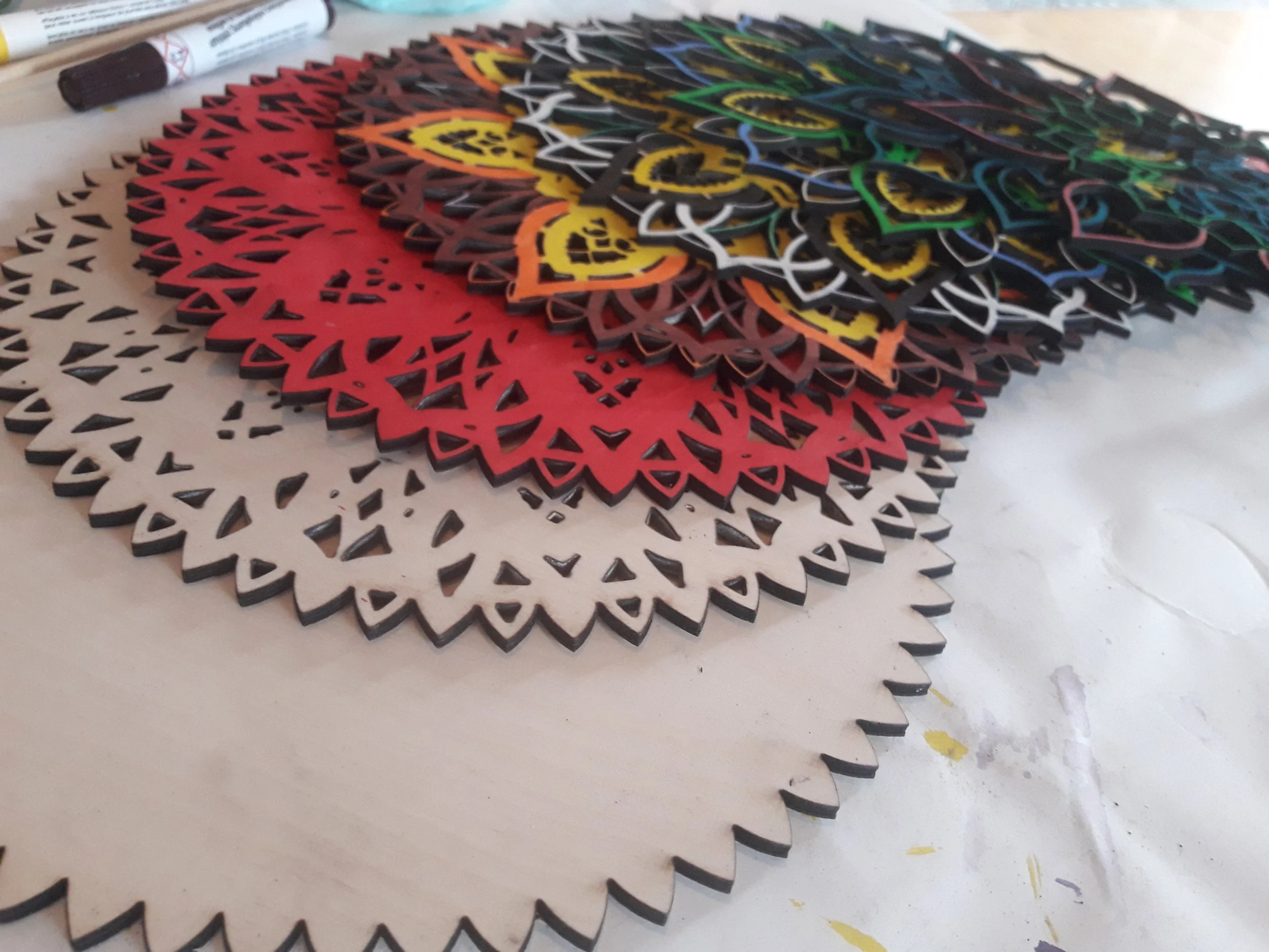 A stack of intricately cut, colorful wooden doilies arranged in overlapping layers. The colors include natural wood, red, yellow, and black, with a marker visible in the background.