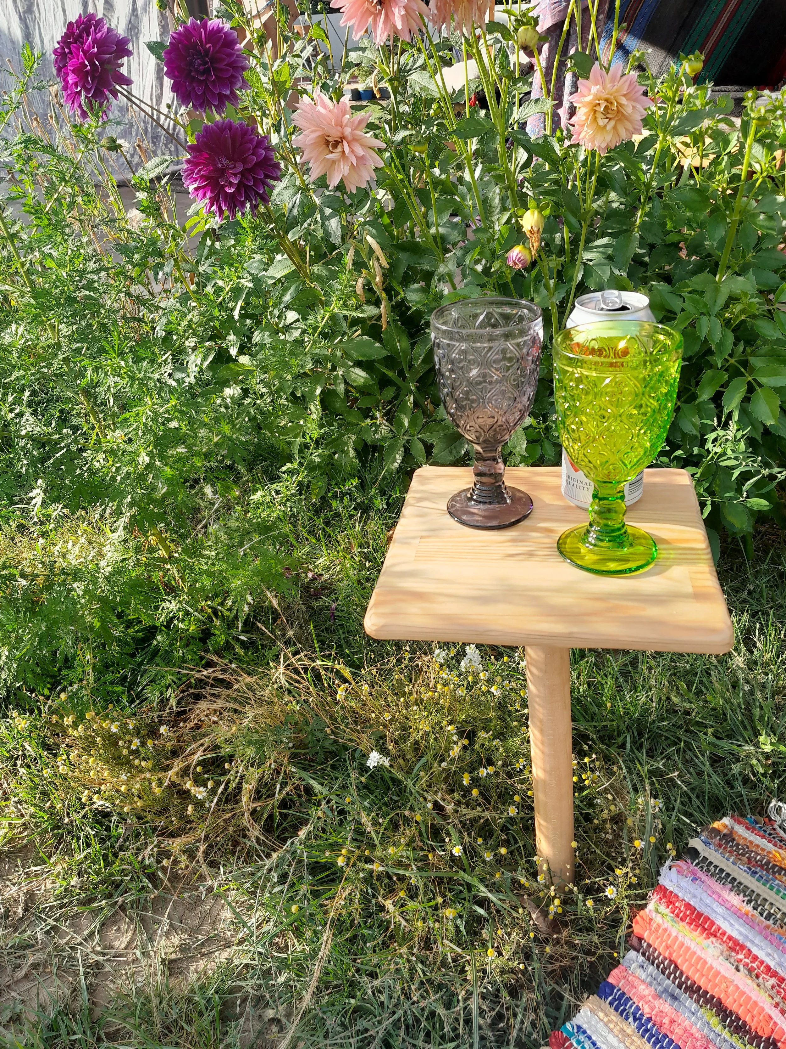 Small wooden table with two vintage-style glasses and a can beside colorful flowers in a garden setting. A striped blanket is partially visible on the ground.