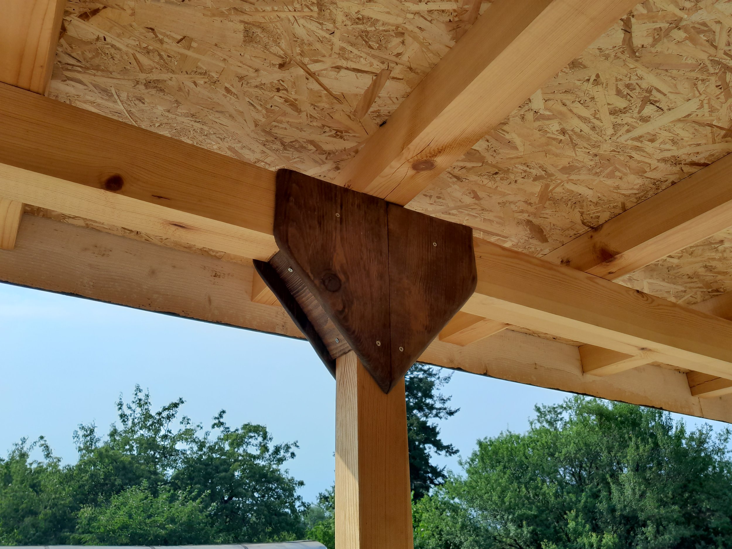 Wooden beams and supports of a partially constructed roof, with oriented strand board (OSB) sheathing and outdoor scenery in the background.