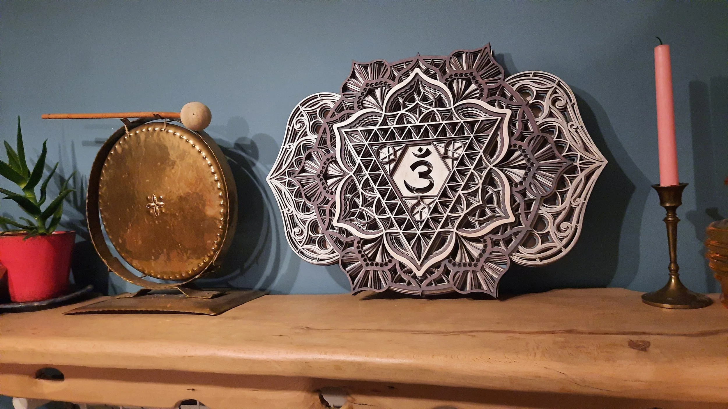 A wooden shelf with a small potted plant in a red pot, a brass gong with a mallet, a decorative wooden mandala design featuring the Om symbol, and a pink candle in a brass holder.