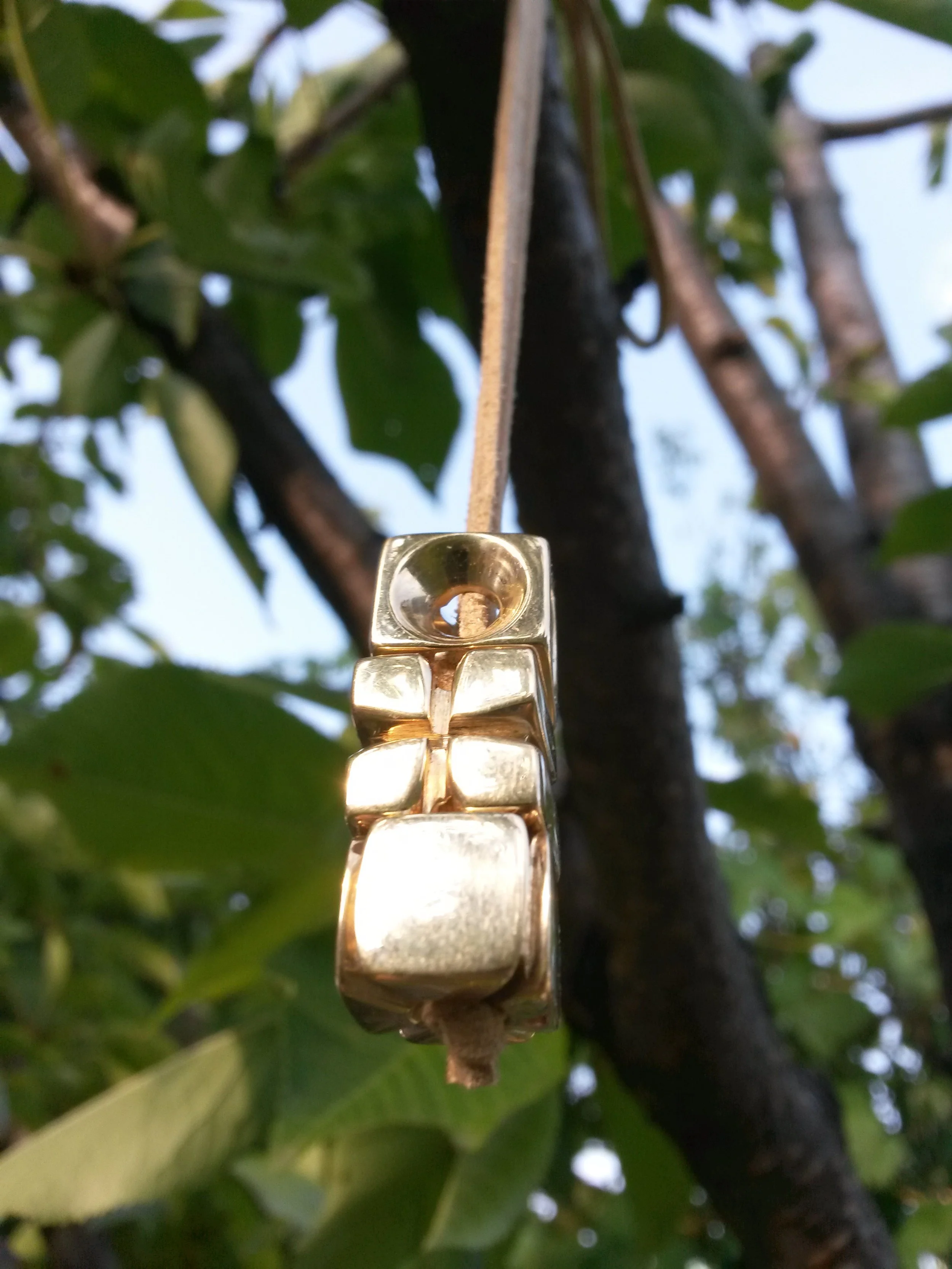 Close-up of a gold geometric pendant hanging on a string against a background of green leaves.