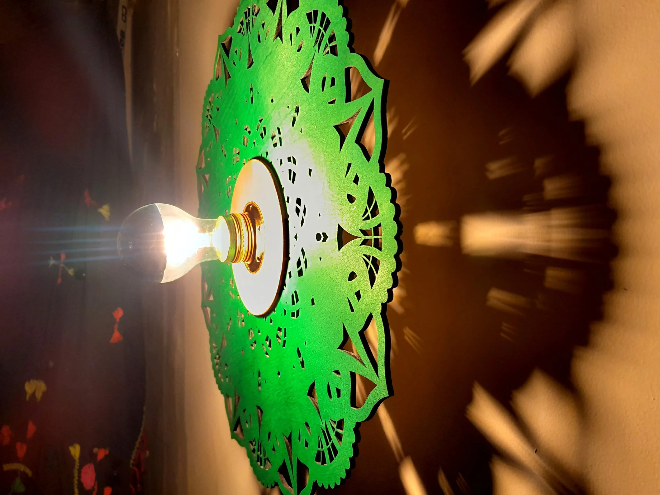 A lit light bulb on a decorative green paper cutout creating intricate shadow patterns on the wall.