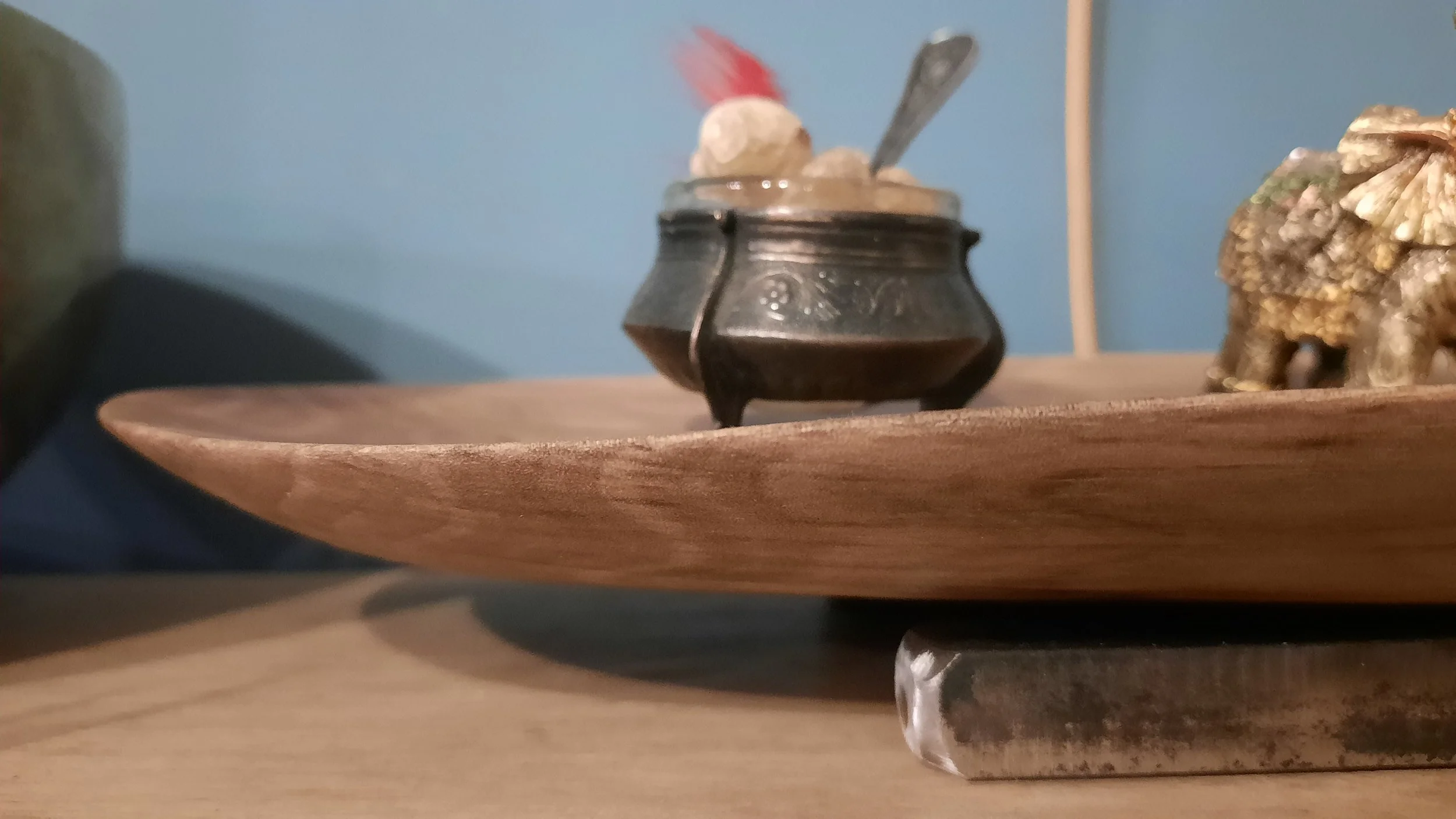 A wooden plate holds a small ornate metal pot containing round objects and a red feather, next to a decorative elephant figurine.