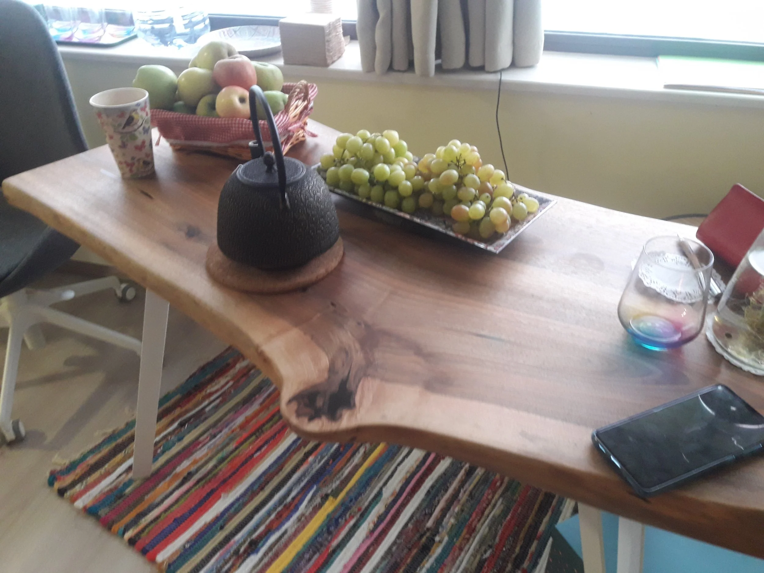 Wooden table with a black teapot, a basket of apples, a plate of grapes, a paper cup, and a smartphone, on a colorful striped rug.