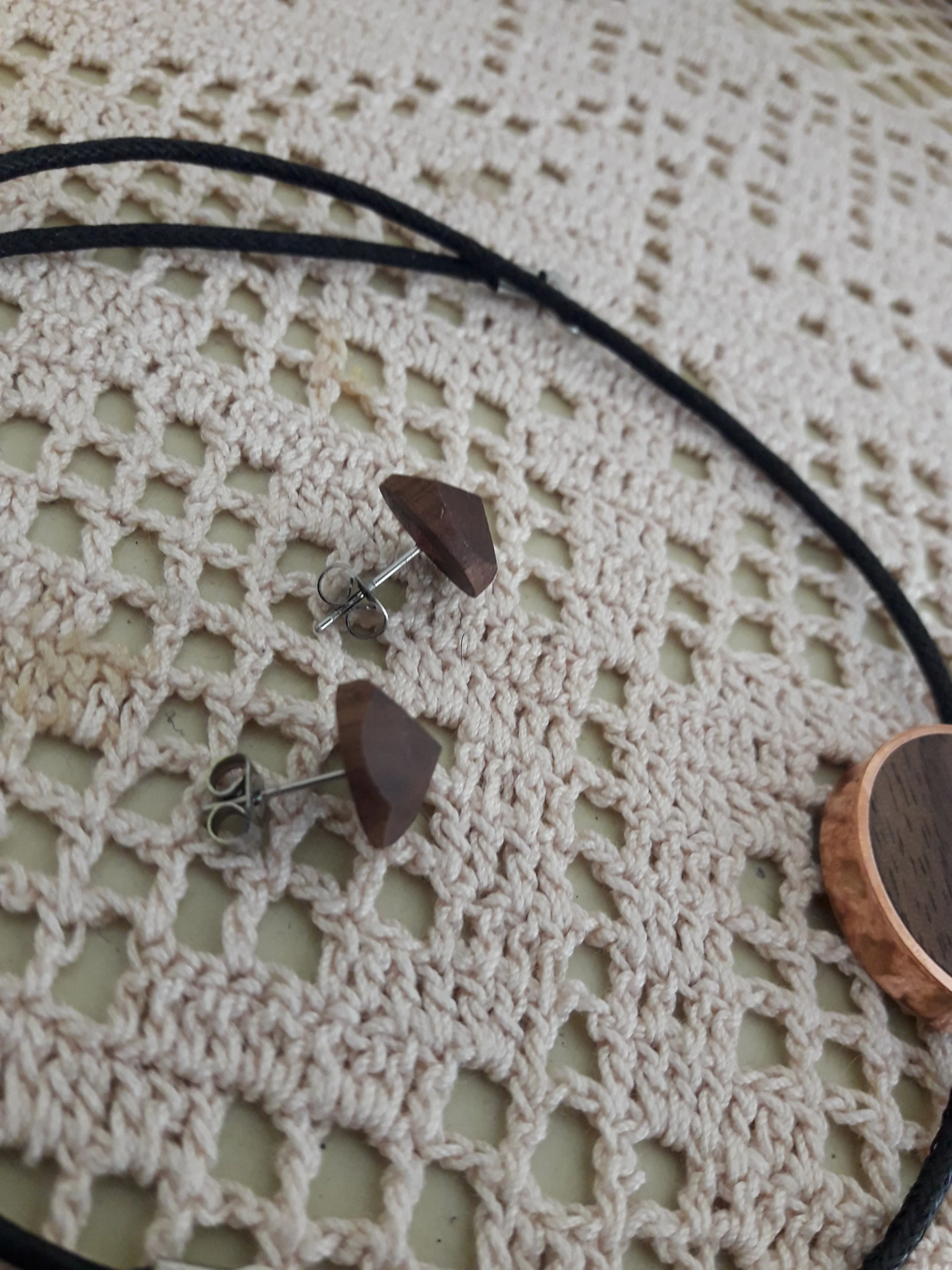 Two wooden stud earrings on a cream-colored crochet fabric next to a black cord necklace and a round wooden accessory.