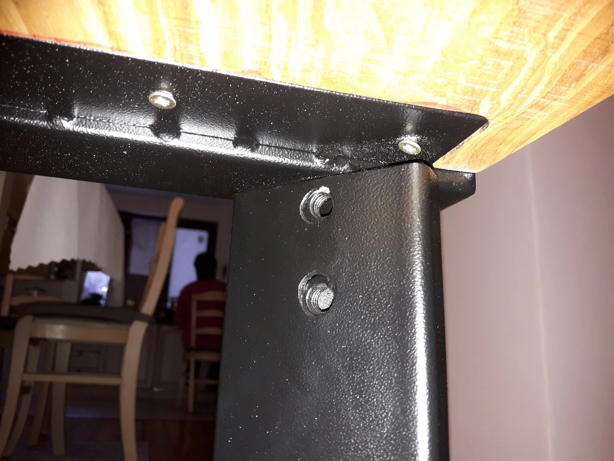 Close-up of a black metal frame attached to a wooden tabletop, with screws visible. Background includes a blurry dining chair and room interior.