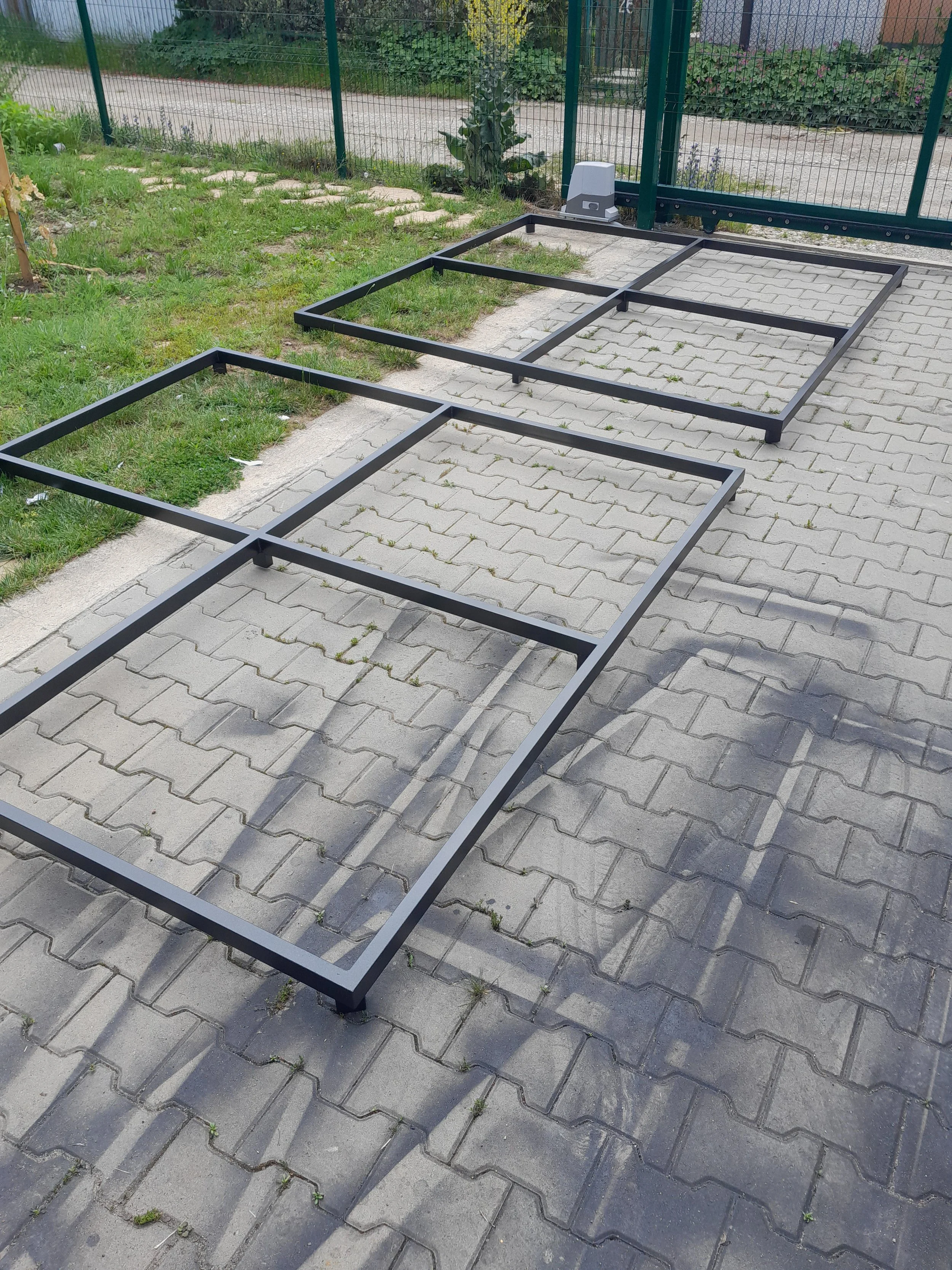 Three metal rectangular frames on a paved outdoor surface next to grass and a fence.