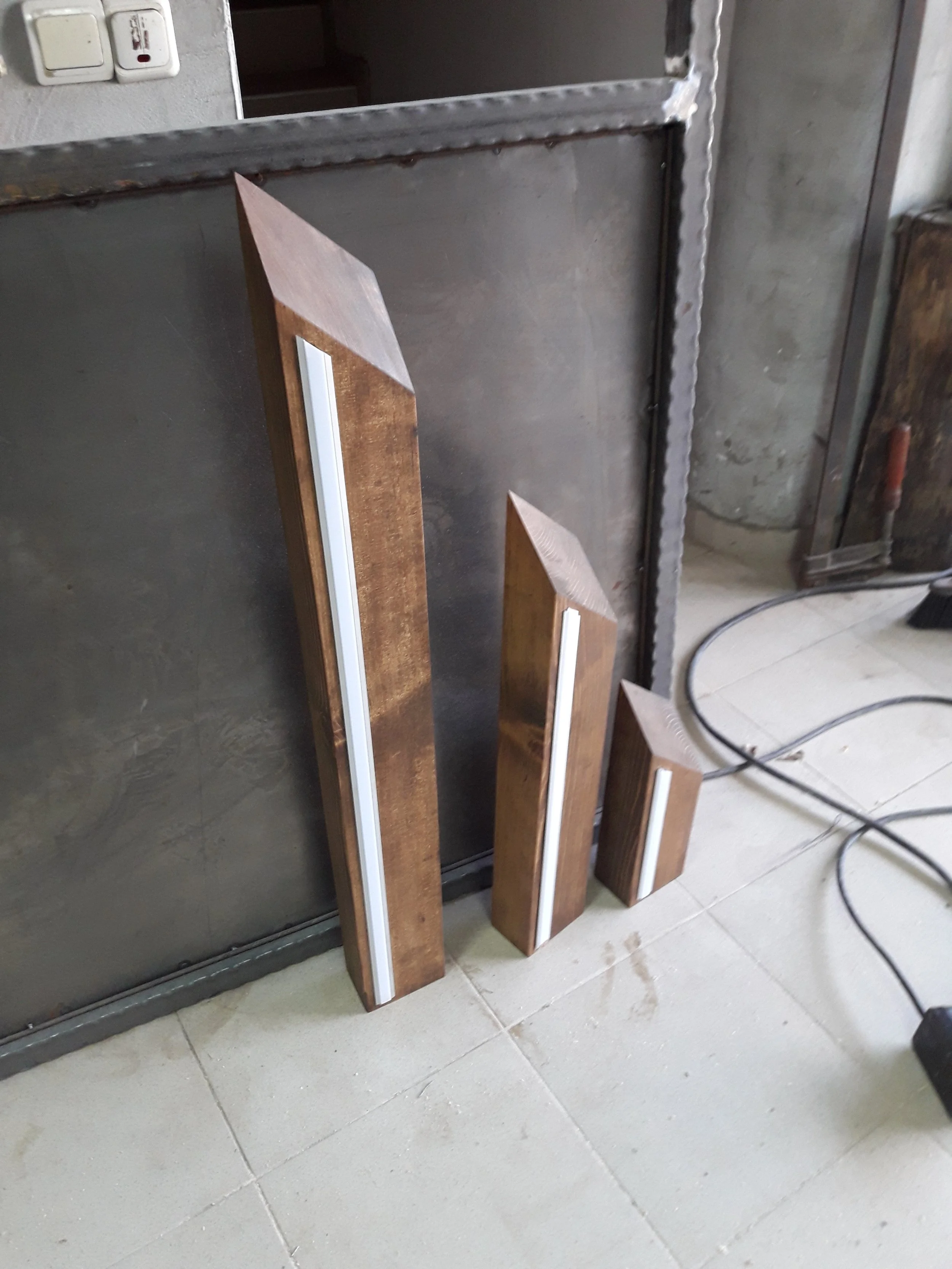 Three wooden LED floor lamps of varying heights with slanted tops and white LED strips, set against a metal wall.