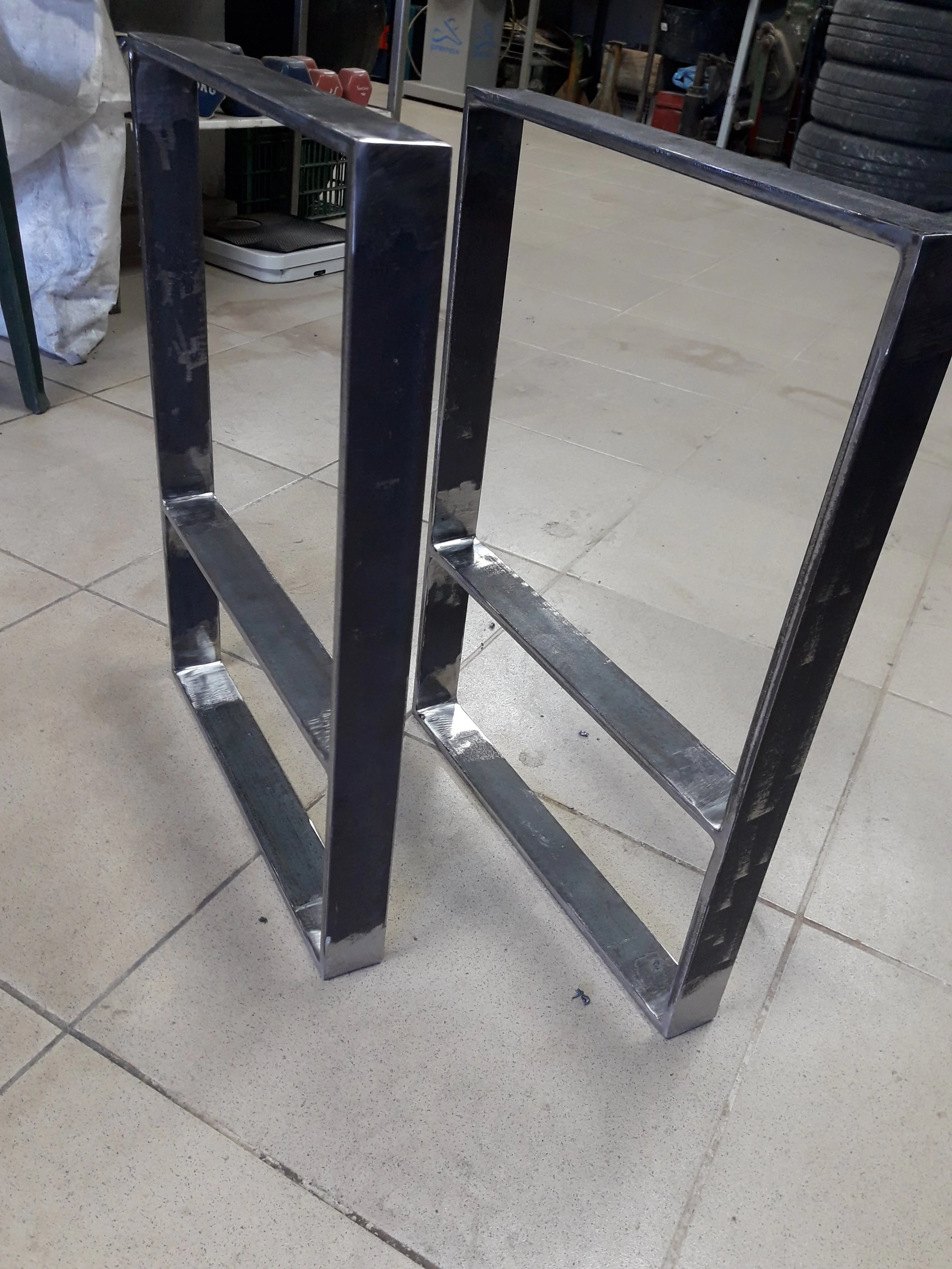 Two rectangular metal frames standing on a tiled floor in a workshop, possibly assembled and welded for furniture or structural support.