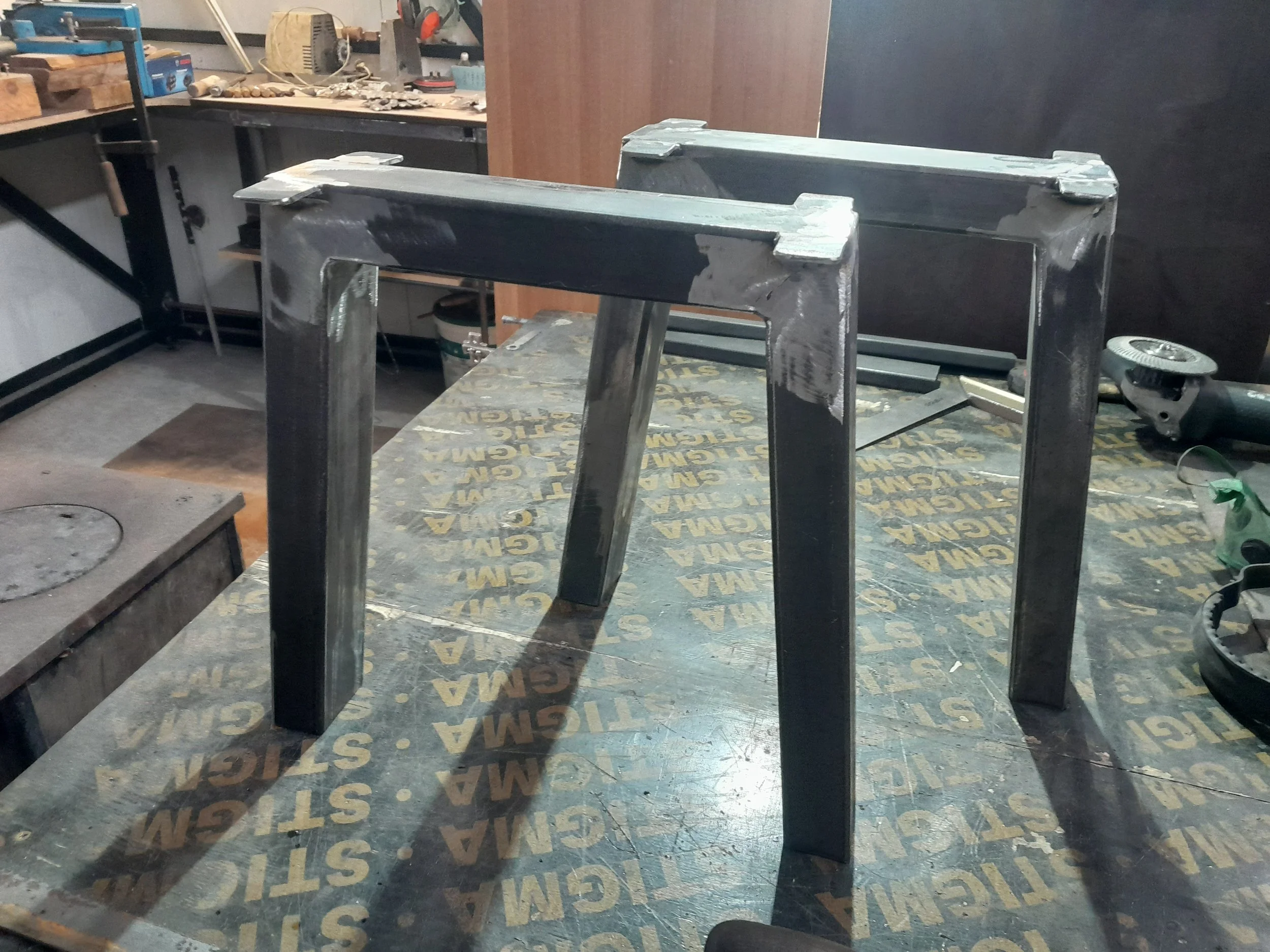 Two metal table legs in a workshop setting, on a workbench with tools and equipment around.