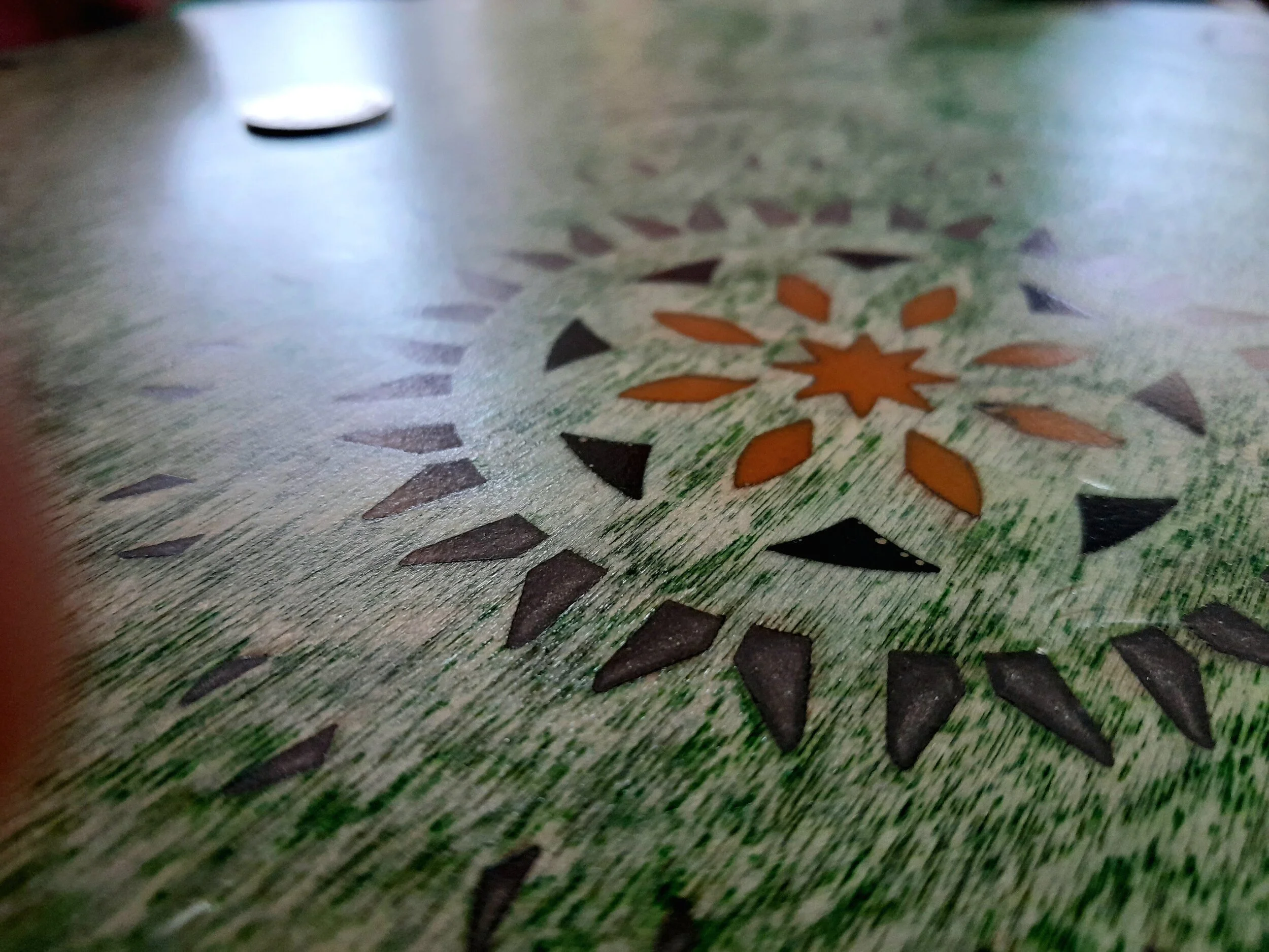 Close-up of a patterned surface with green coloring, featuring orange and black geometric shapes arranged in a circular design. A blurred coin is visible in the background.