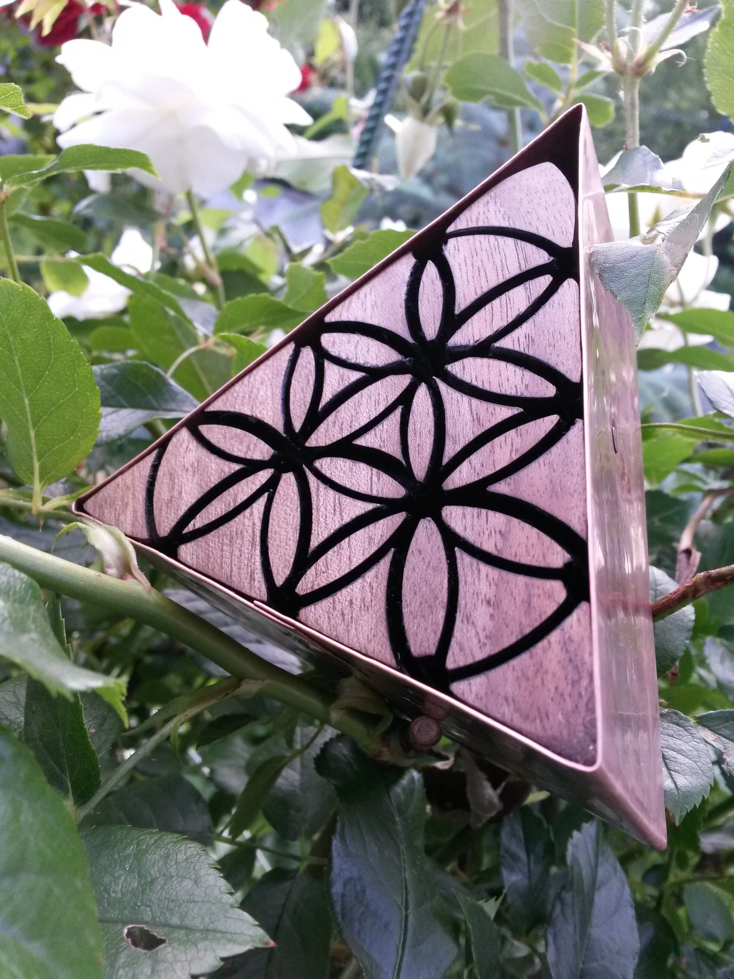 Triangular wooden art piece with geometric pattern amidst green foliage and white flowers.