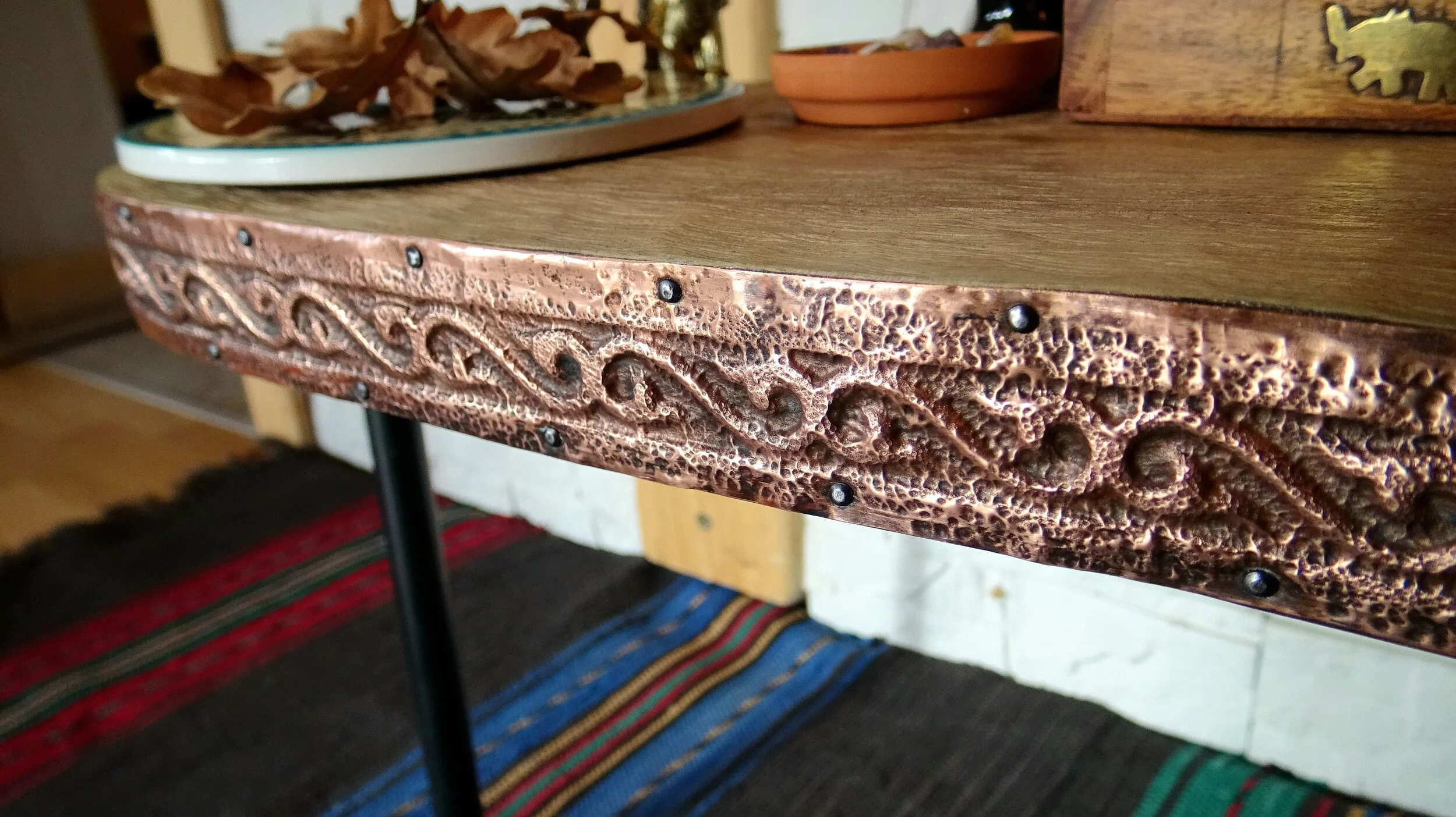 Close-up of a table edge with decorative metal trim and rustic finish, displaying intricate patterns. The table surface holds decorative items like a ceramic dish and a potted plant. A vibrant woven rug is partially visible beneath the table.