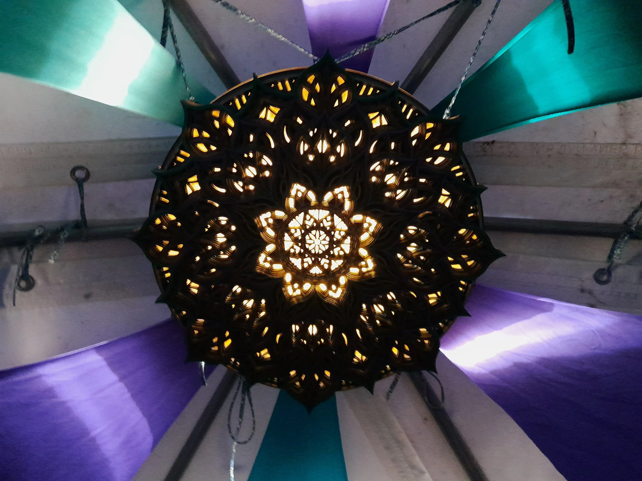 Decorative chandelier with intricate patterns, hanging from a ceiling with colorful fabric drapes in green, purple, and white.