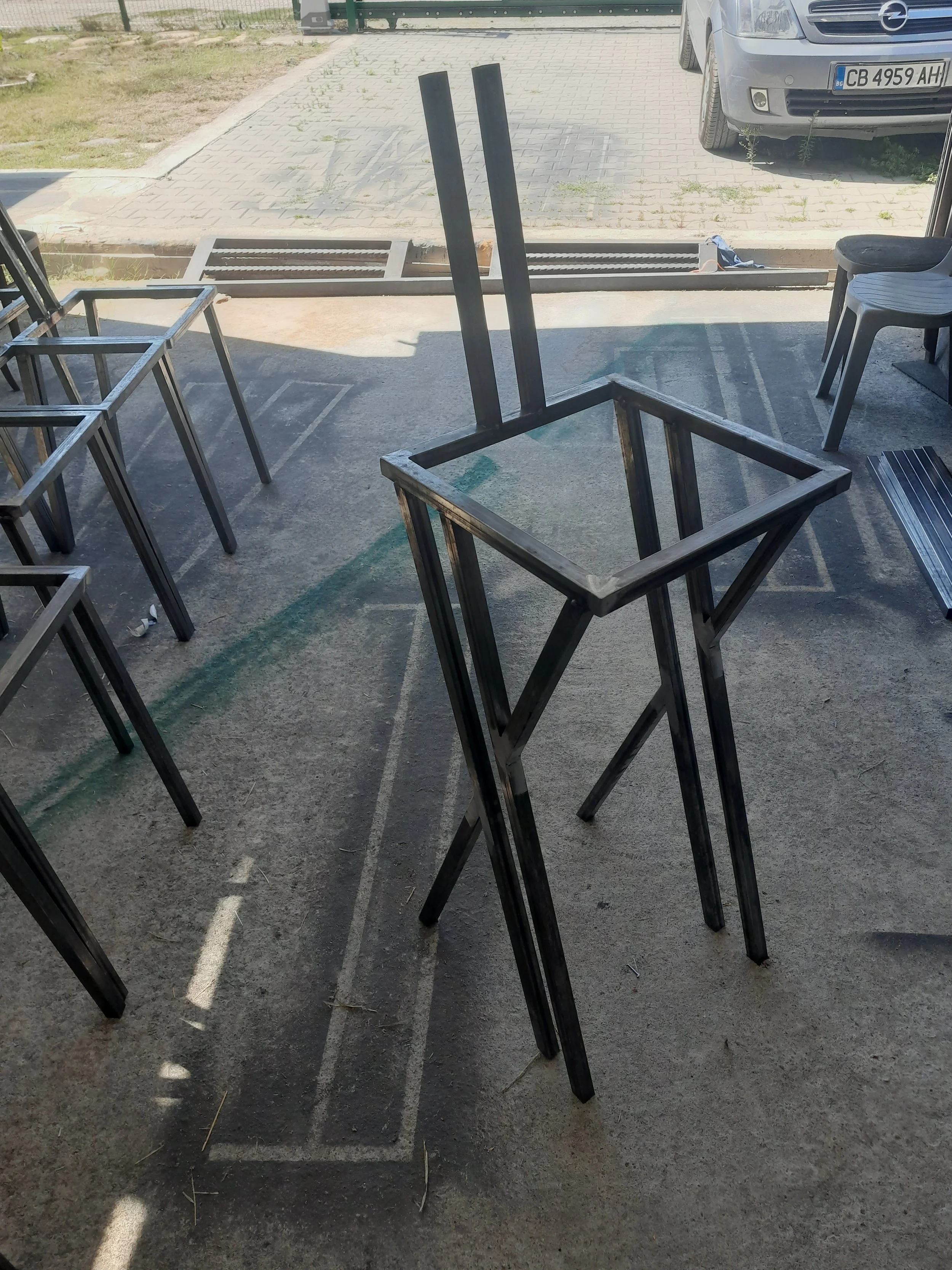 Metal frame for a tall chair with back support, placed on a concrete floor, surrounded by other similar frames, near an outdoor space with a car in the background.